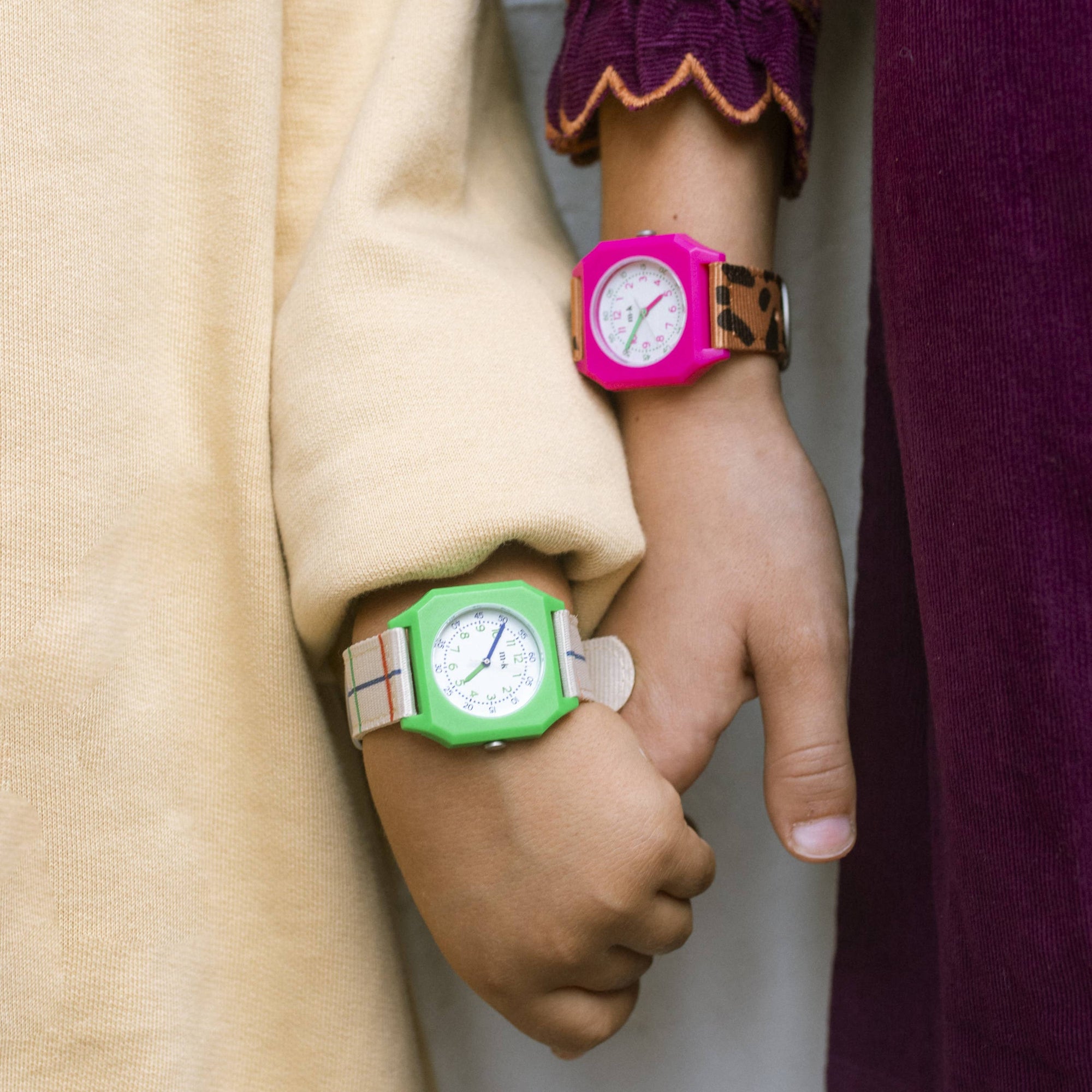 Two hands holding each other, one wearing a green watch and the other a pink watch, against a plain background.
