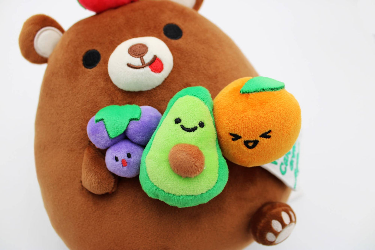 A close-up of a brown plush bear holding three small fruit and vegetable plush characters: purple grapes, a green avocado, and an orange with a green leaf.