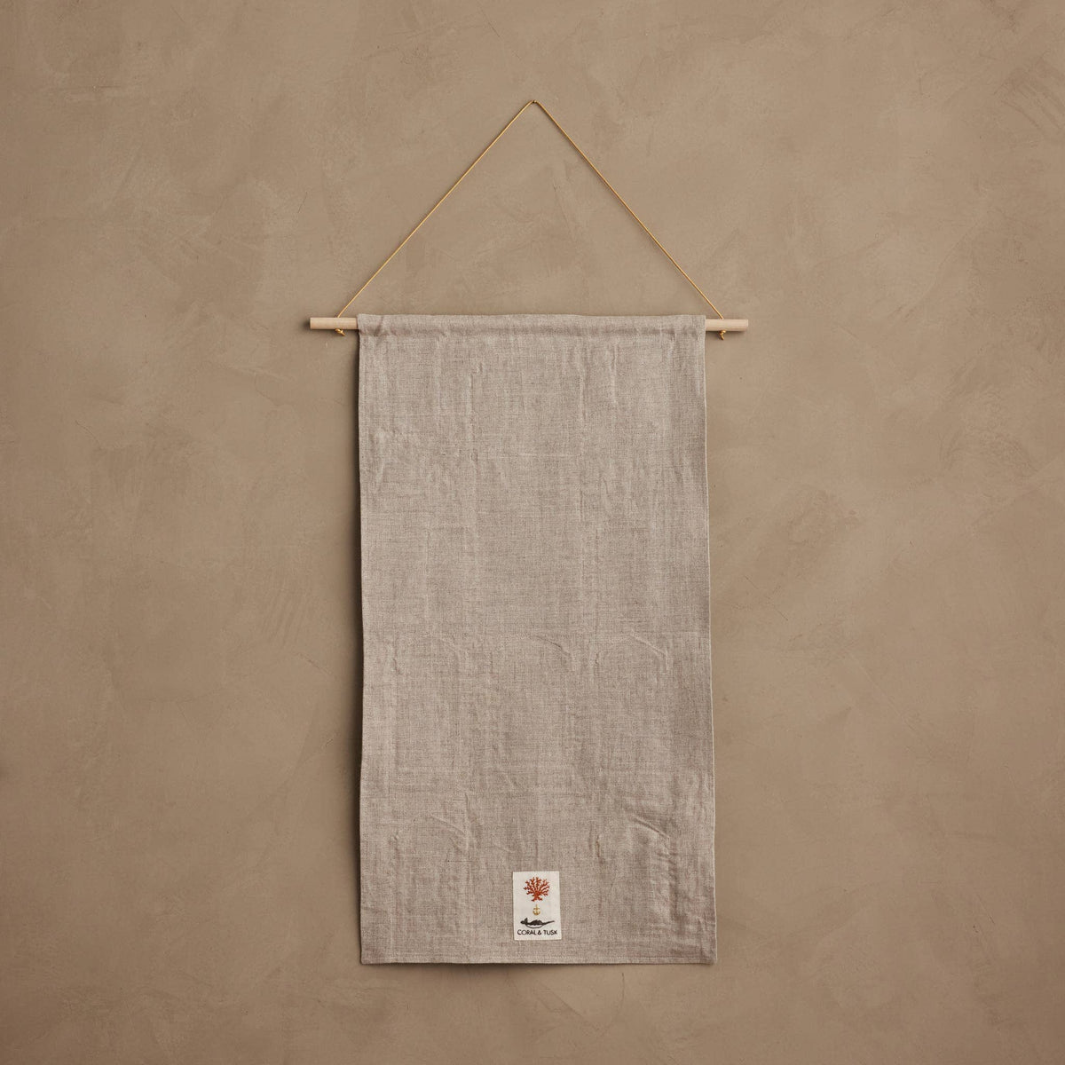 A light beige linen wall hanging towel suspended from a wooden dowel with a string, set against a textured beige plaster wall. A small white brand label is visible at the bottom.