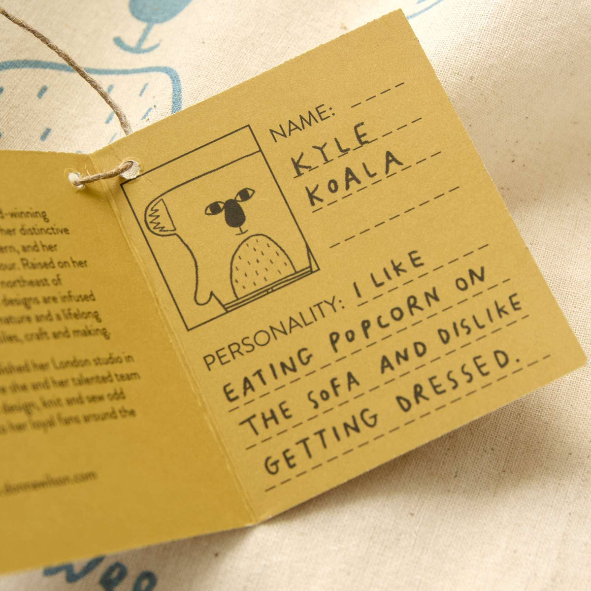 A close-up of a handmade paper passport-style card with a string loop, printed with a cartoon koala and text fields like name and personality. The card is shown partially opened on a light fabric background.