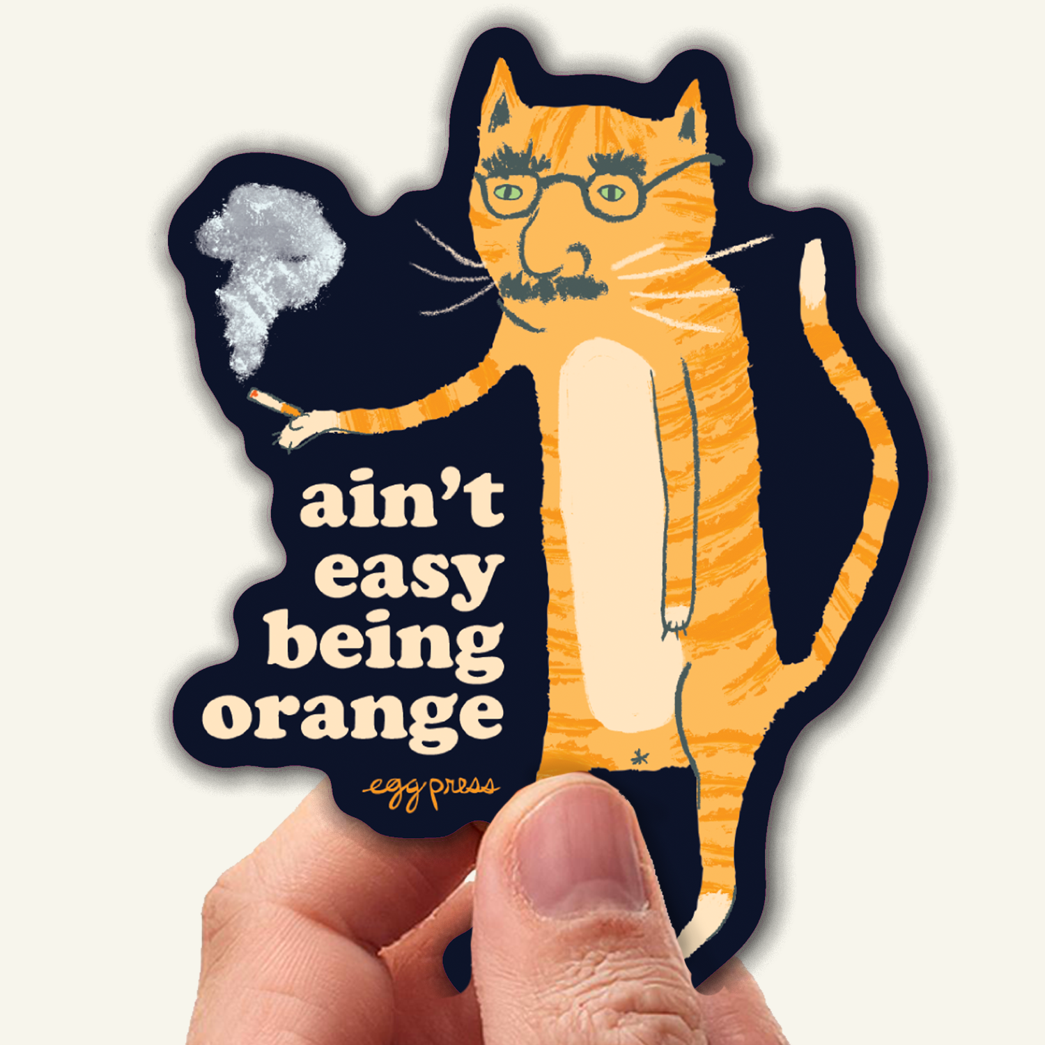 Sticker of a cat with glasses and the text 'ain't easy being orange' held by a hand.