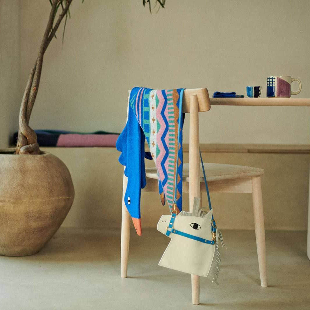 A styled interior scene with a light wood dining table, chair, and large ceramic planter. A colorful patterned scarf and a blue-and-white plush toy are draped over the chair, alongside a small white horse-shaped handbag with blue trim.