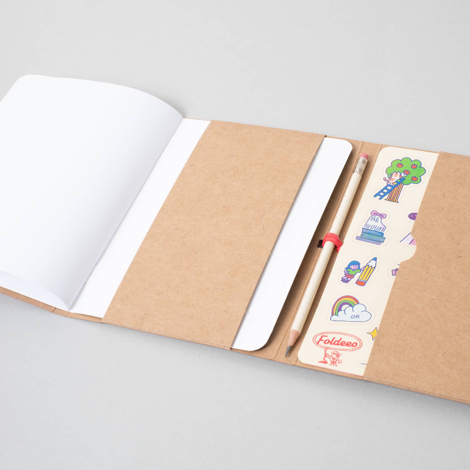 A kraft paper notebook with an open blank white page on the left and a closed white page on the right, secured with a red elastic band. A pencil and a decorative sticker strip with school-themed illustrations are tucked into the right side pocket.