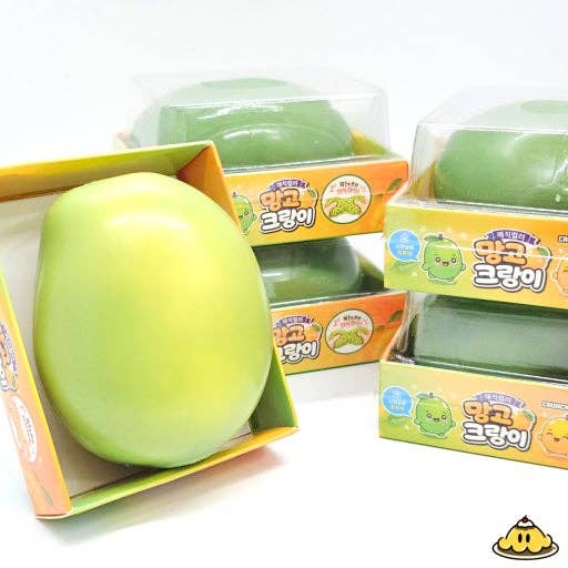A green mango-shaped squishy toy shown open in its packaging, with several similar packaged toys stacked in the background on a white surface.