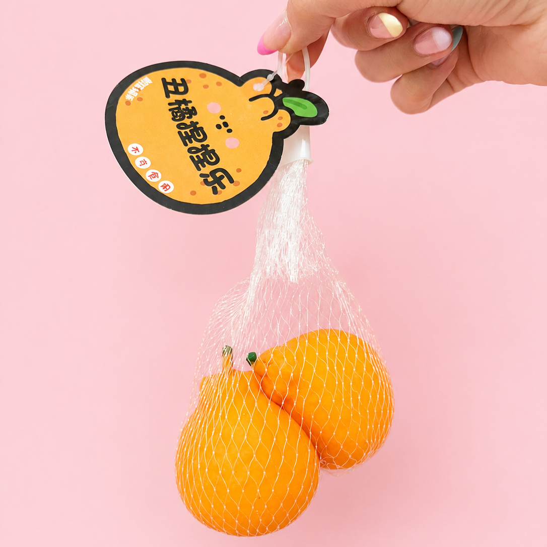 A hand holds a white mesh bag containing two bright oranges against a solid pink background. The bag has a decorative yellow tag with Chinese text and a cartoon  orange illustration.