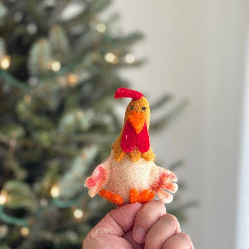 Small felt rooster ornament held in front of a decorated Christmas tree.