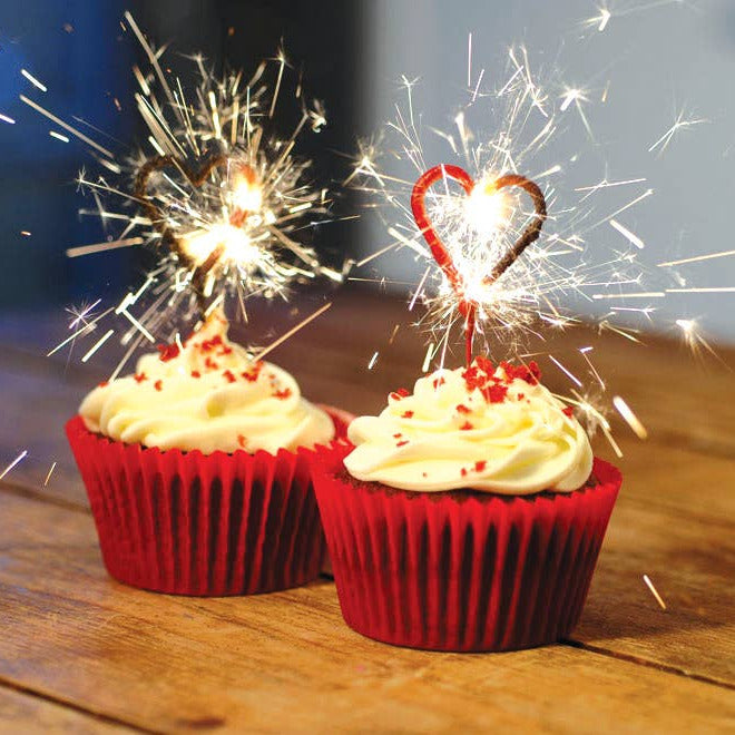 Two cupcakes with red wrappers and white frosting, each topped with a lit sparkler, on a wooden surface.