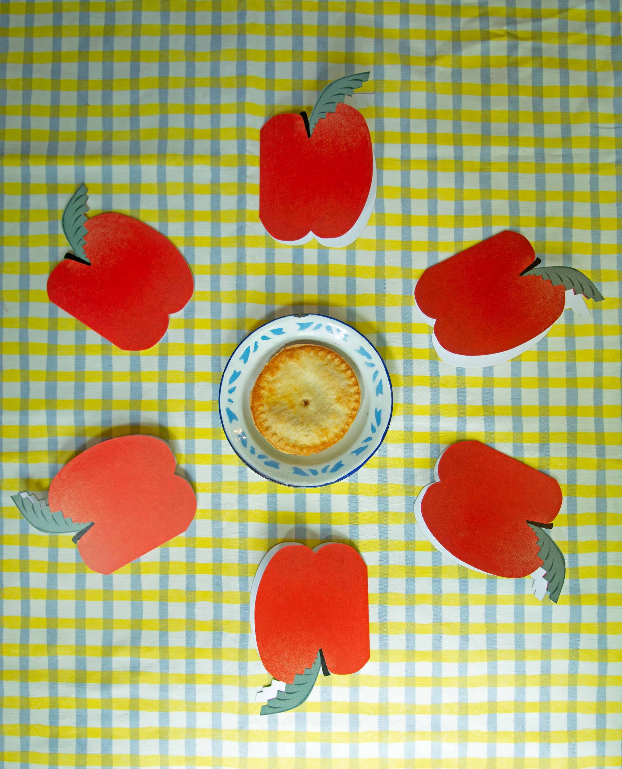 Red apple-shaped cookies around a small round dish on a yellow and blue checkered tablecloth.