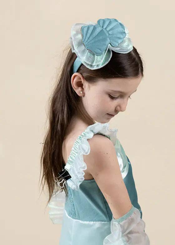 A young girl with long brown hair is shown from the side and back, wearing a light blue costume dress with ruffled trim, sheer white arm cuffs, and a matching light blue shell-shaped hair bow headband against a solid beige background.