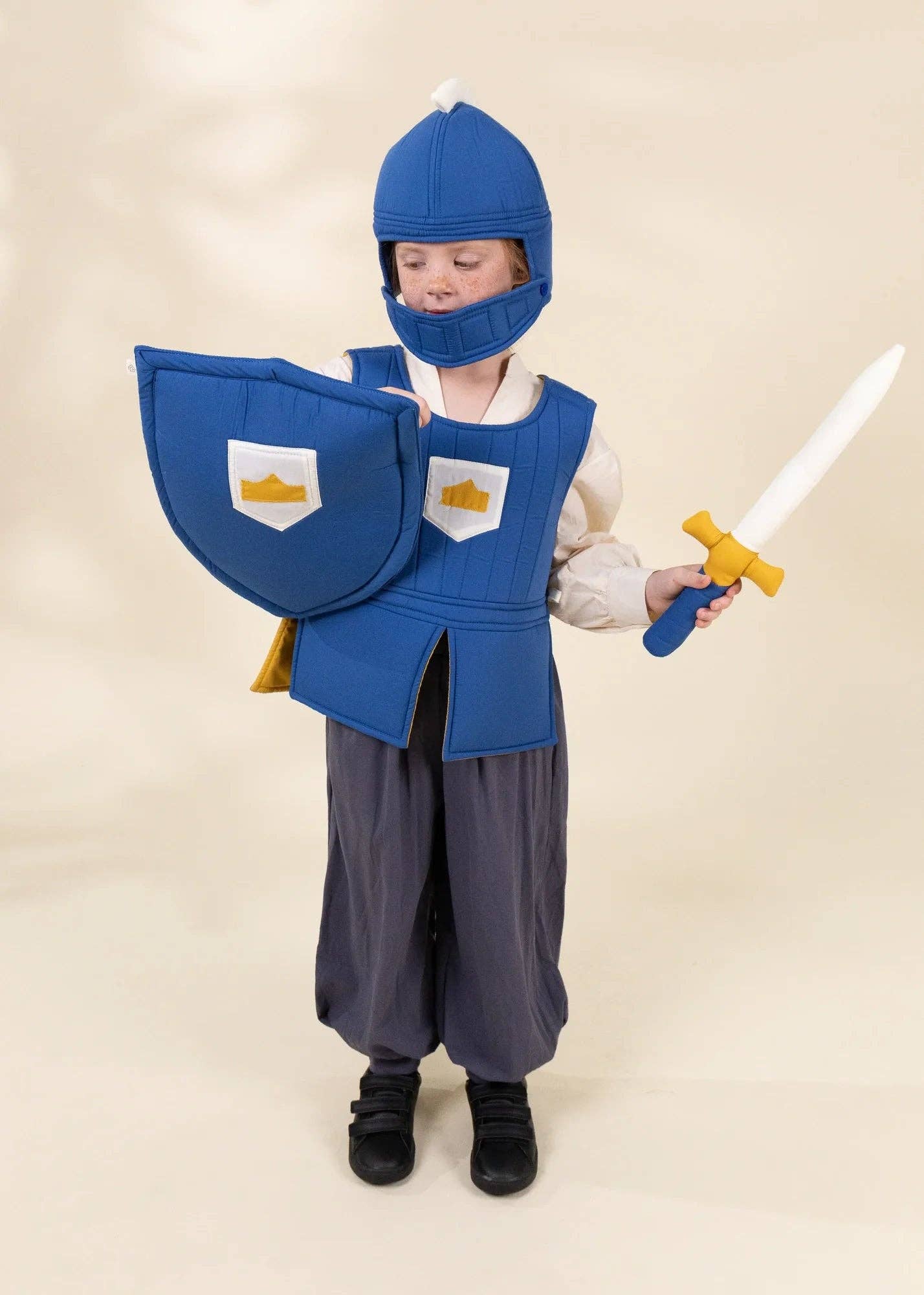 A child wearing a medieval knight costume stands against a plain beige studio background, holding a toy sword and wearing a blue helmet, blue tunic, gray pants, and black shoes.