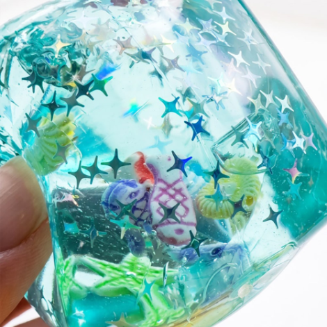 A close-up of a hand holding a translucent blue-green cube-shaped decorative item filled with glitter, star-shaped foil, and small multicolored decorative pieces.