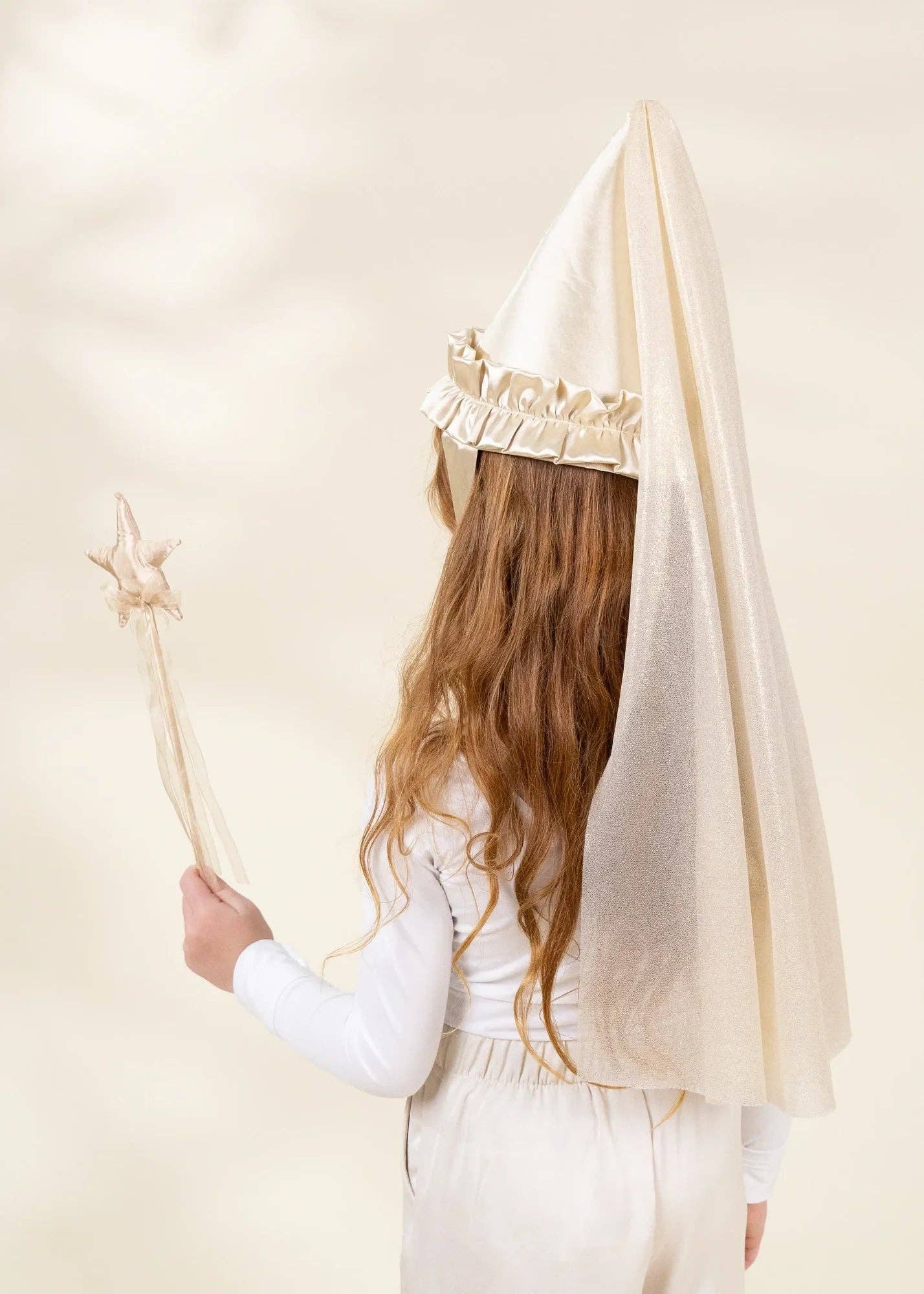A child seen from behind wearing a cream-colored pointed head covering with a ruffled trim and a long wavy light brown hair, holding a matching wand against a soft beige background.