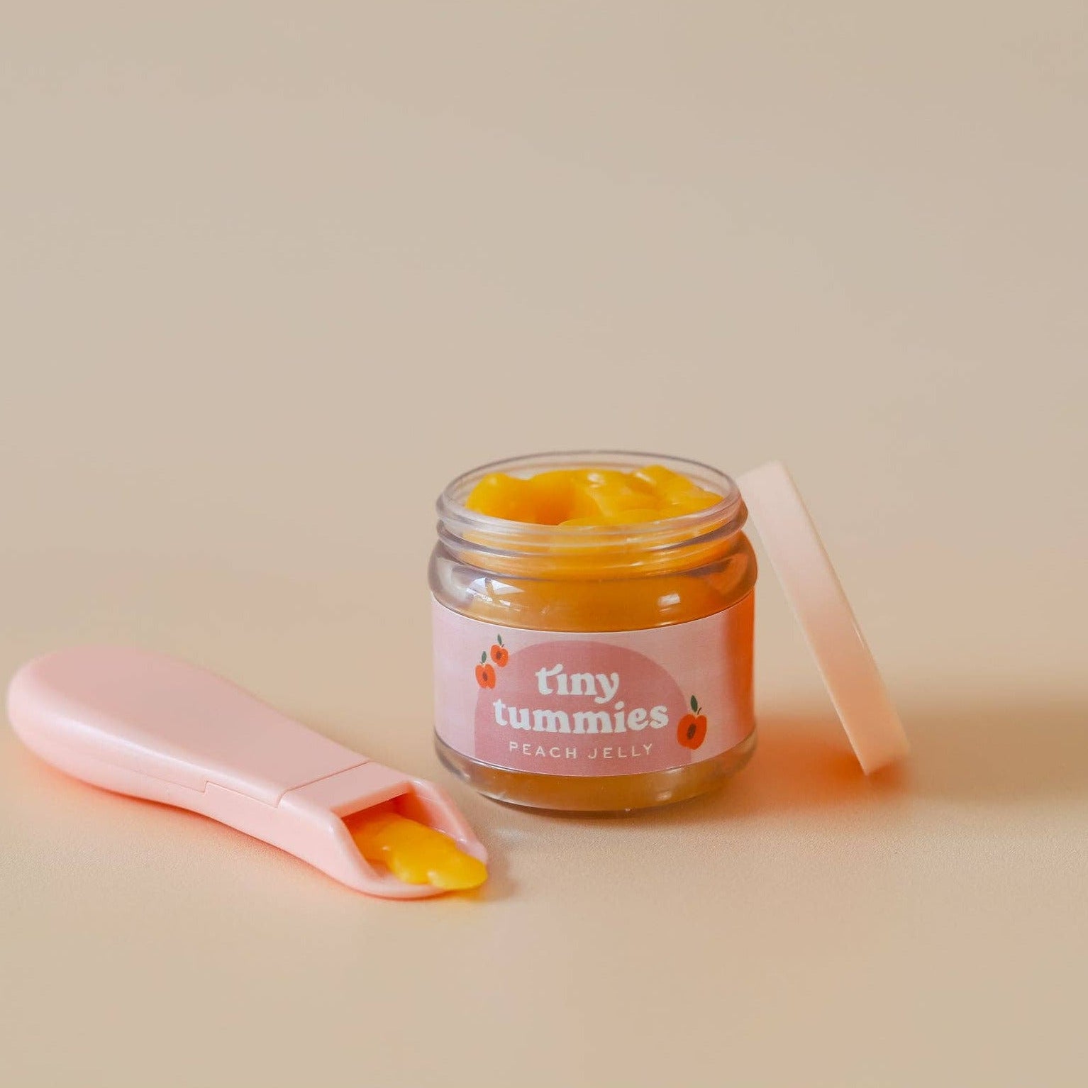 Jar of 'tiny tummies' baby food with a pink spoon on a beige background