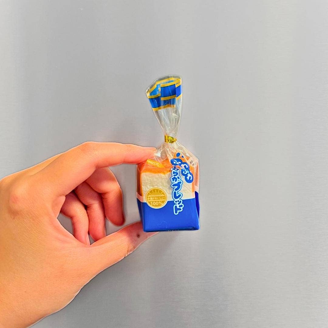 A hand holding a miniature loaf of bread-shaped item wrapped in clear cellophane with blue and yellow packaging, set against a plain gray background.