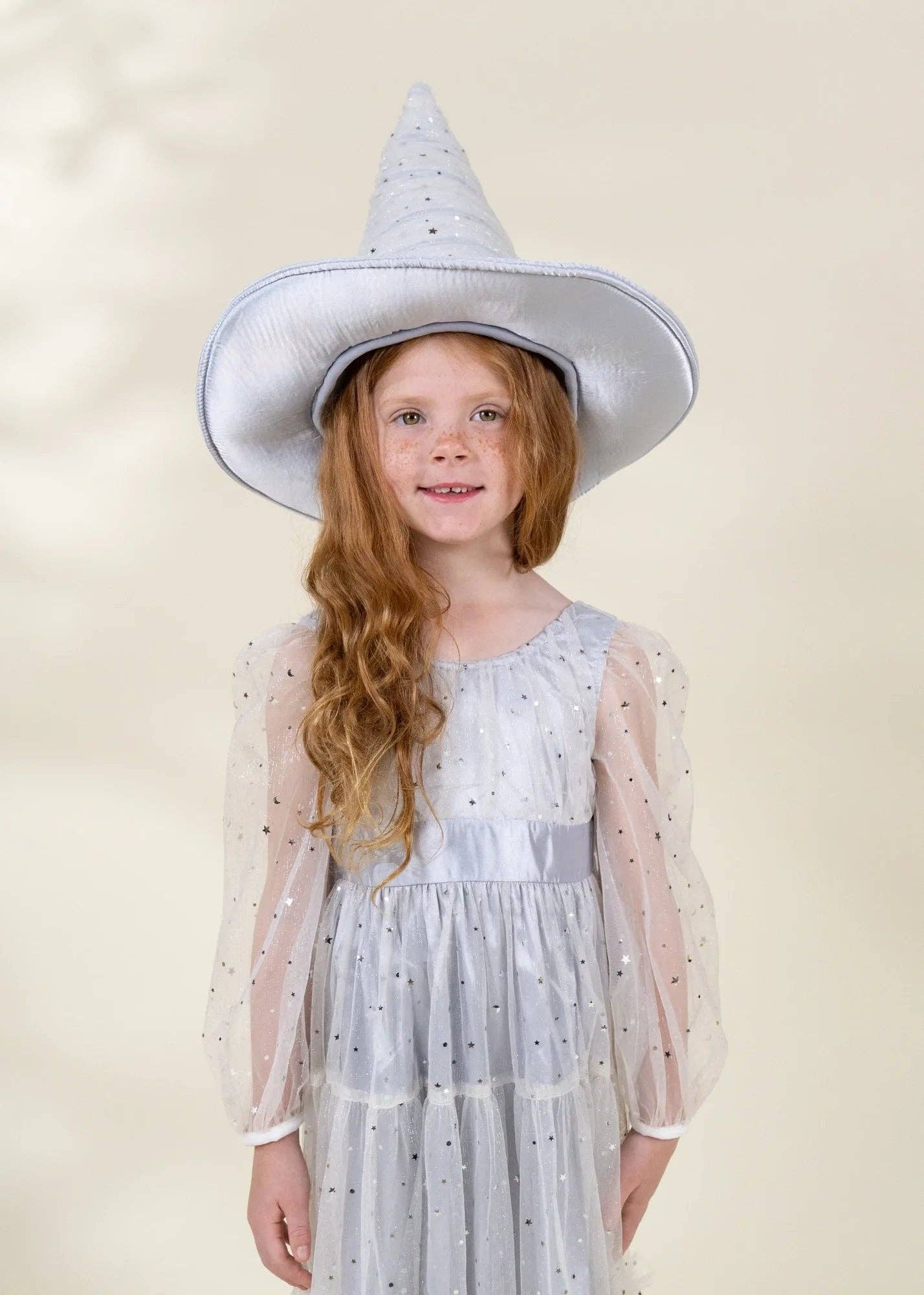 A young girl with long wavy red hair wearing a white witch hat and a white tulle dress with silver polka dots, standing against a soft beige background.