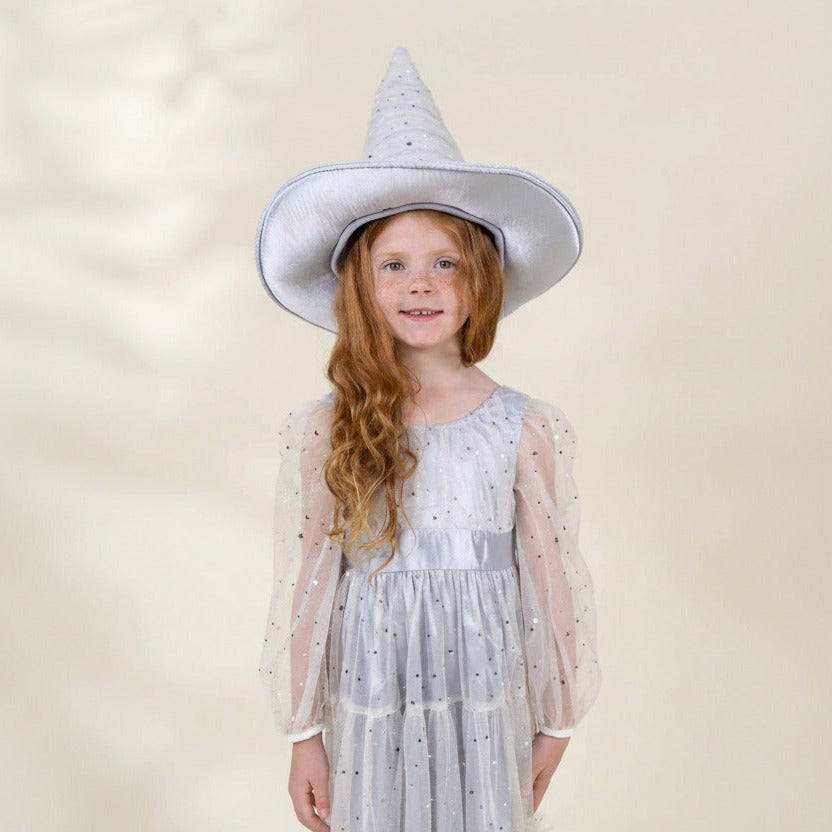 A young girl with long wavy red hair wearing a white witch hat and a white tulle dress with silver polka dots, standing against a soft beige background.