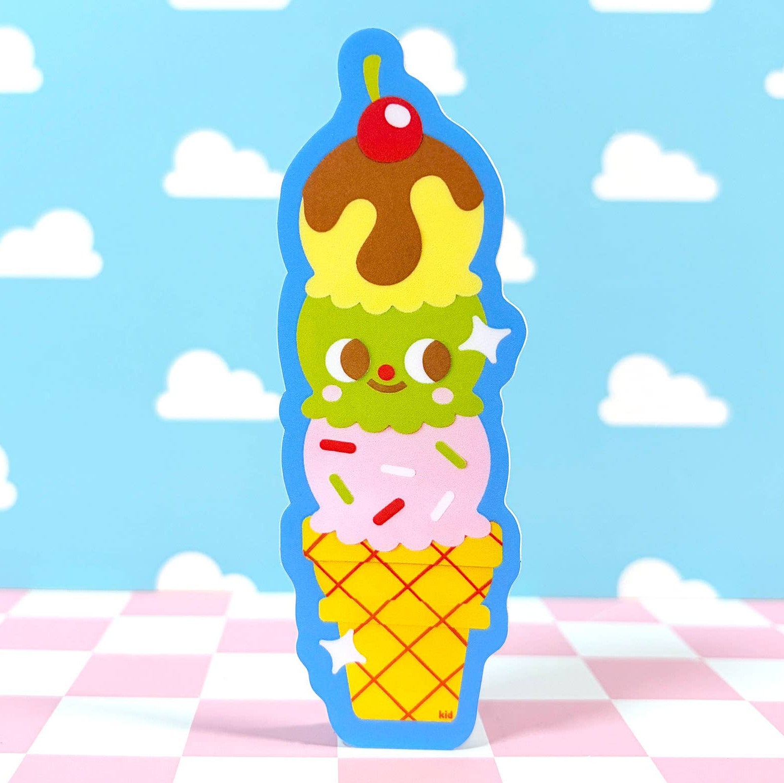 Ice cream cone-shaped plush toy with cartoon face on a blue background with white clouds and pink checkered tablecloth.
