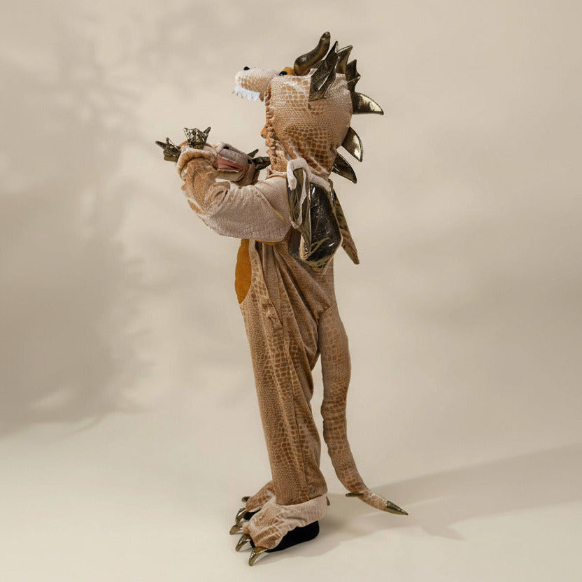 A person wearing a full-body dragon costume stands against a plain beige studio background. The costume includes a textured tan and brown body, metallic gold spikes along the head and back, a long tail, clawed feet, and small wing-like pieces on the shoulders.