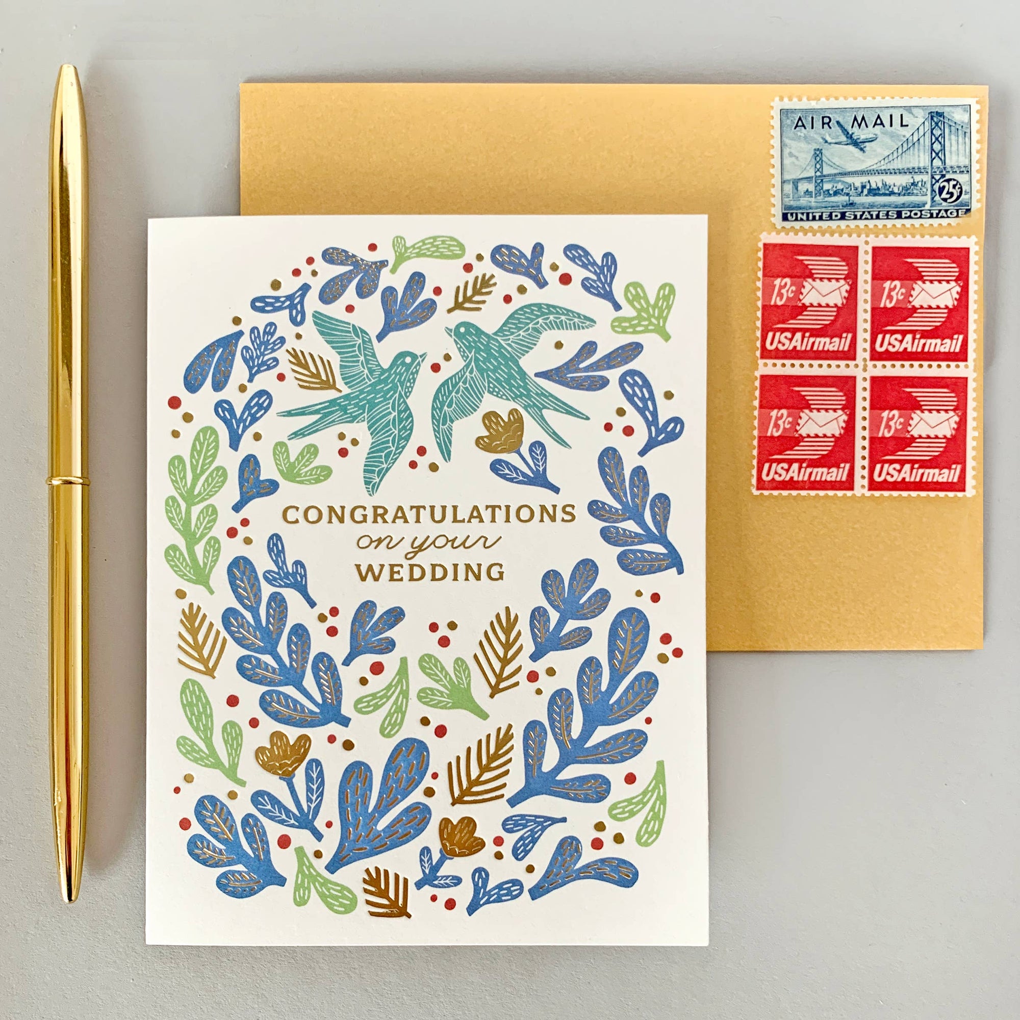Wedding card with decorative birds and leaves, gold pen, and U.S. postage stamps on a gray background