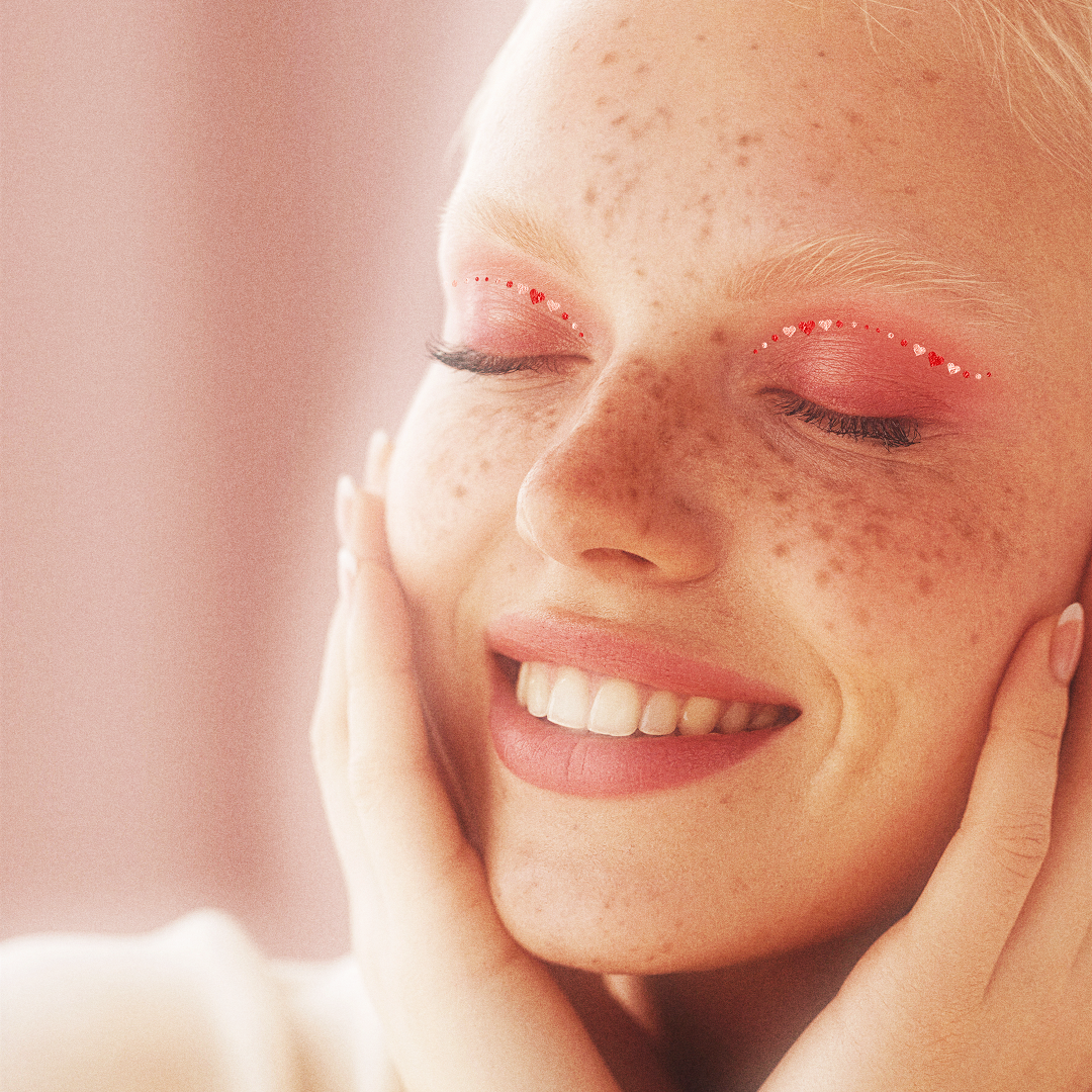 Close-up of a woman with freckles smiling softly against a pink background