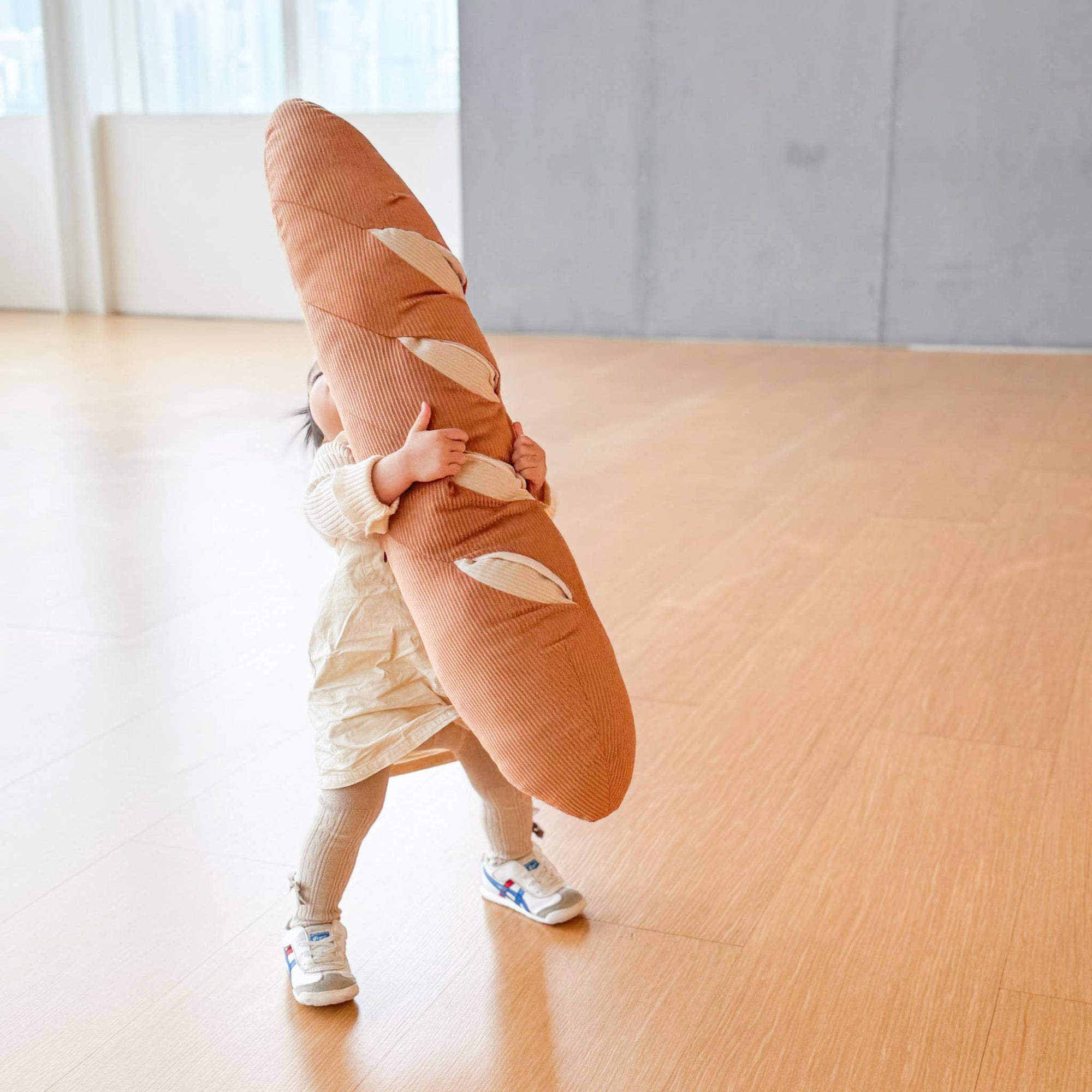 Child holding a large bread-shaped pillow on a wooden floor