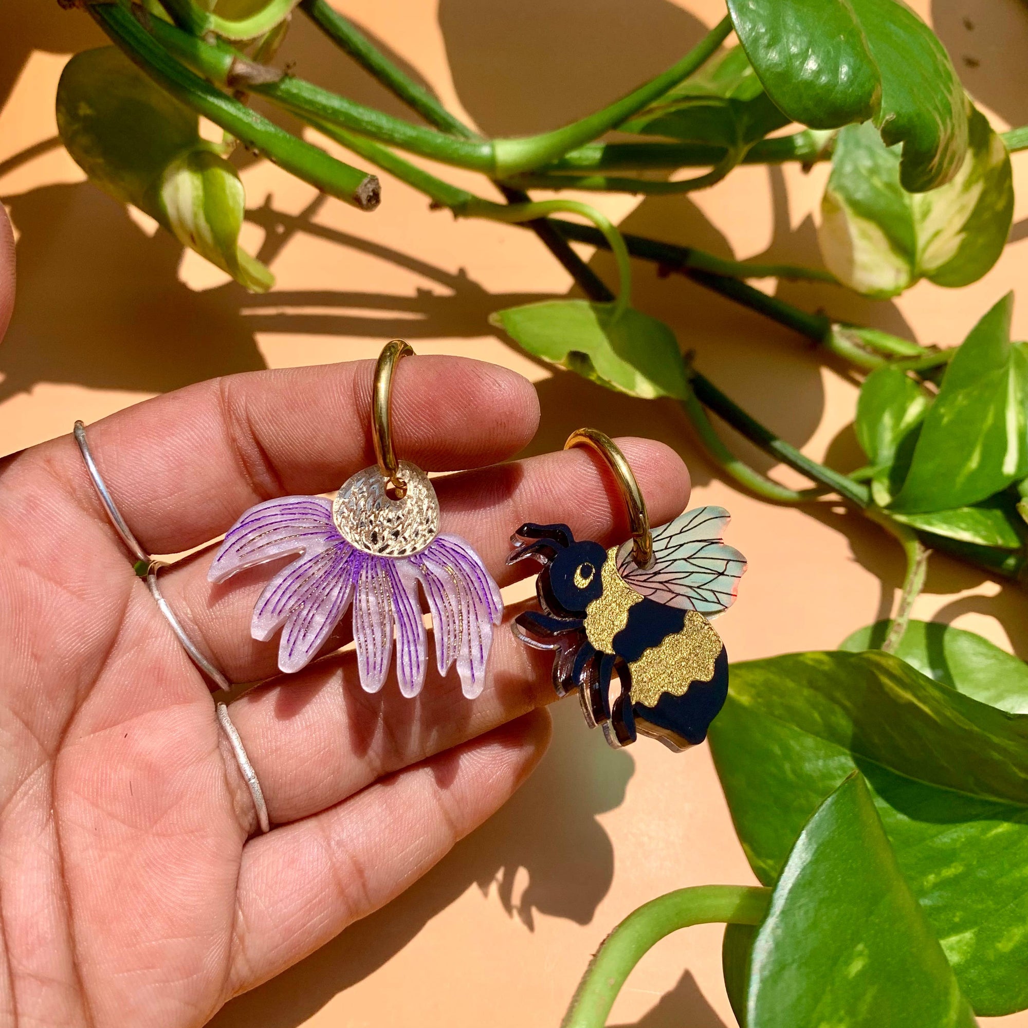 Hand holding two earrings: a purple flower and a bee design, with green leaves in the background.