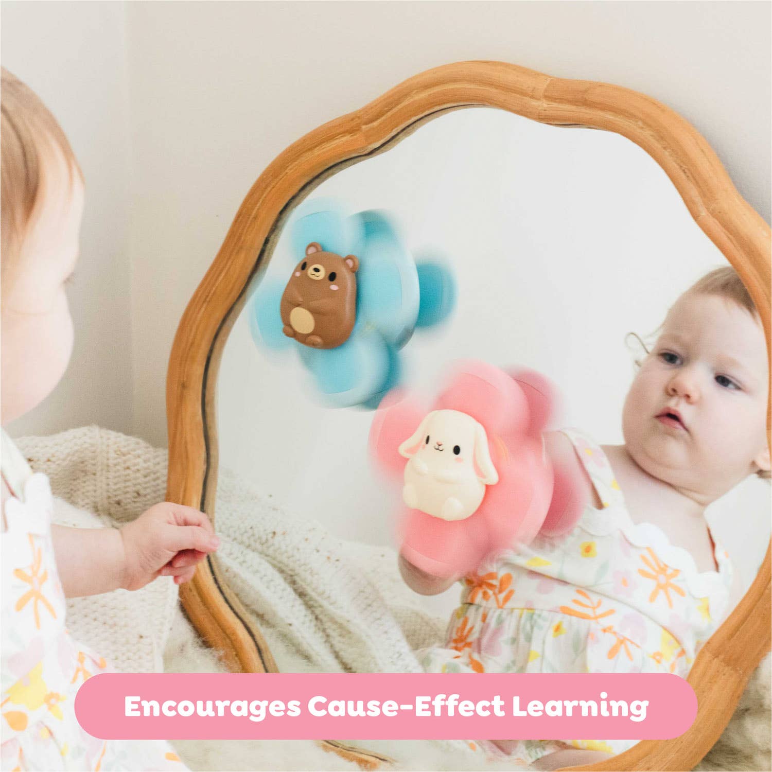 Baby looking at itself in a mirror with two soft toys, one brown bear and one pink sheep, held by another child.