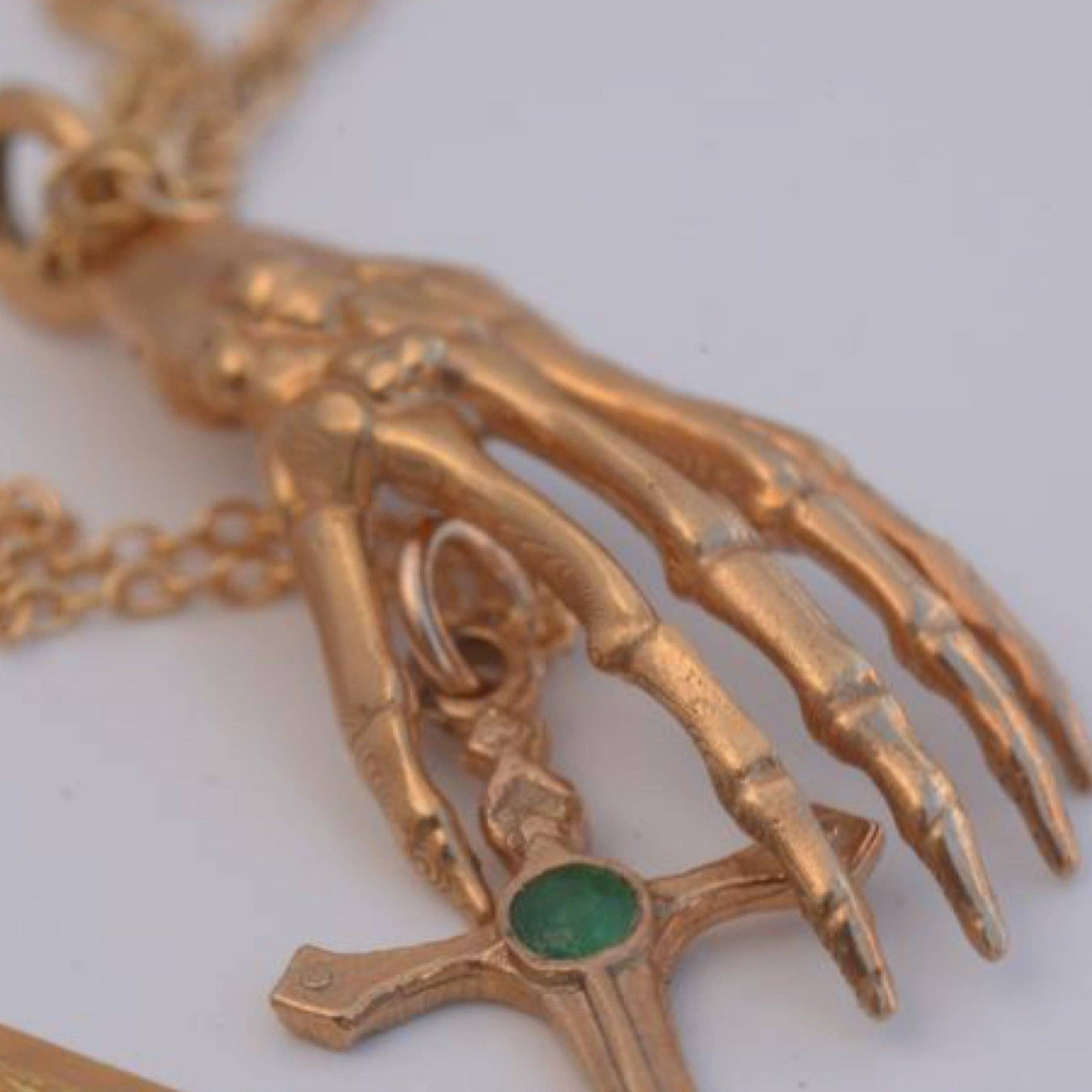 Gold skeleton hand pendant with green gemstone on a white background