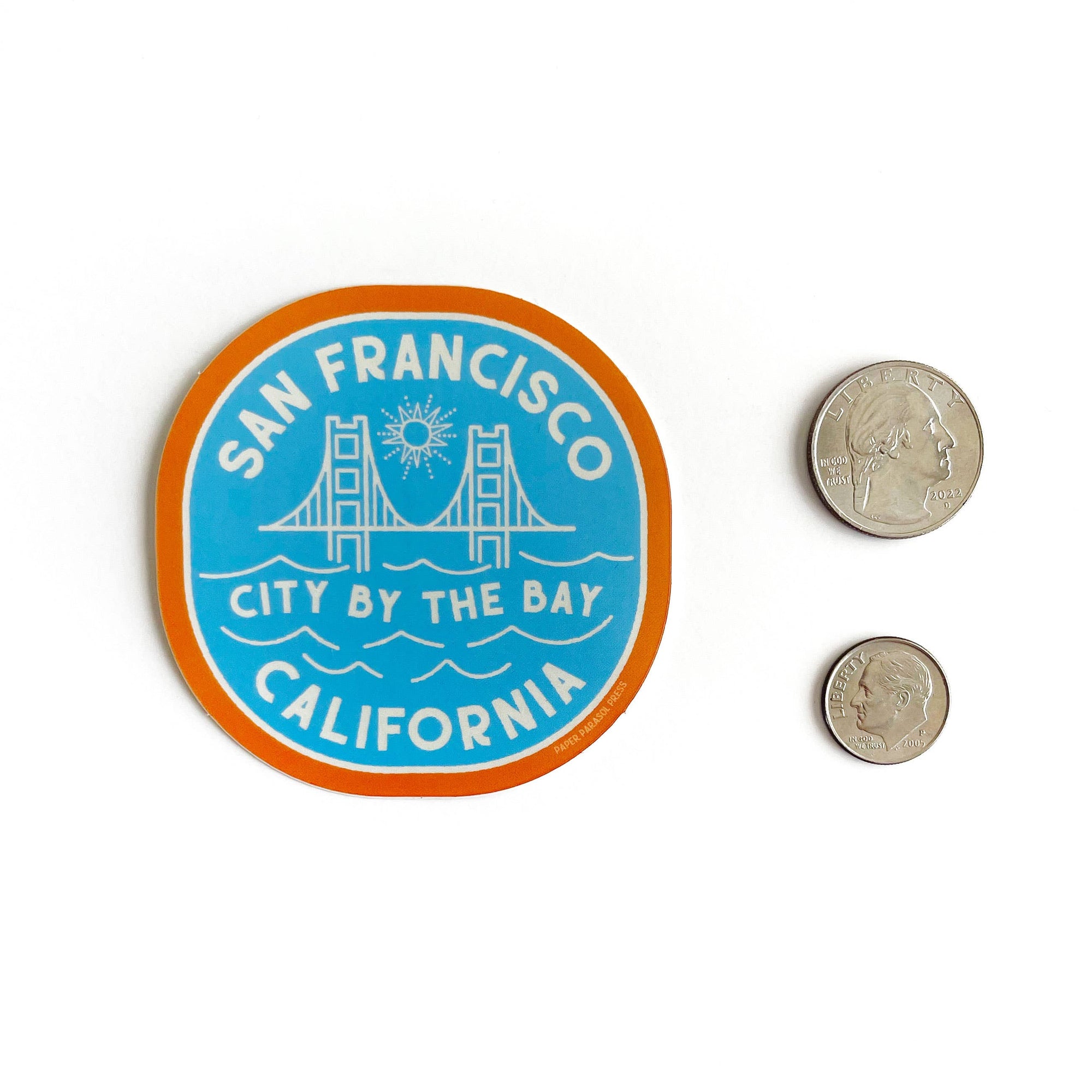 Round sticker with 'San Francisco, City by the Bay, California' text and a bridge graphic, next to two coins for size comparison.