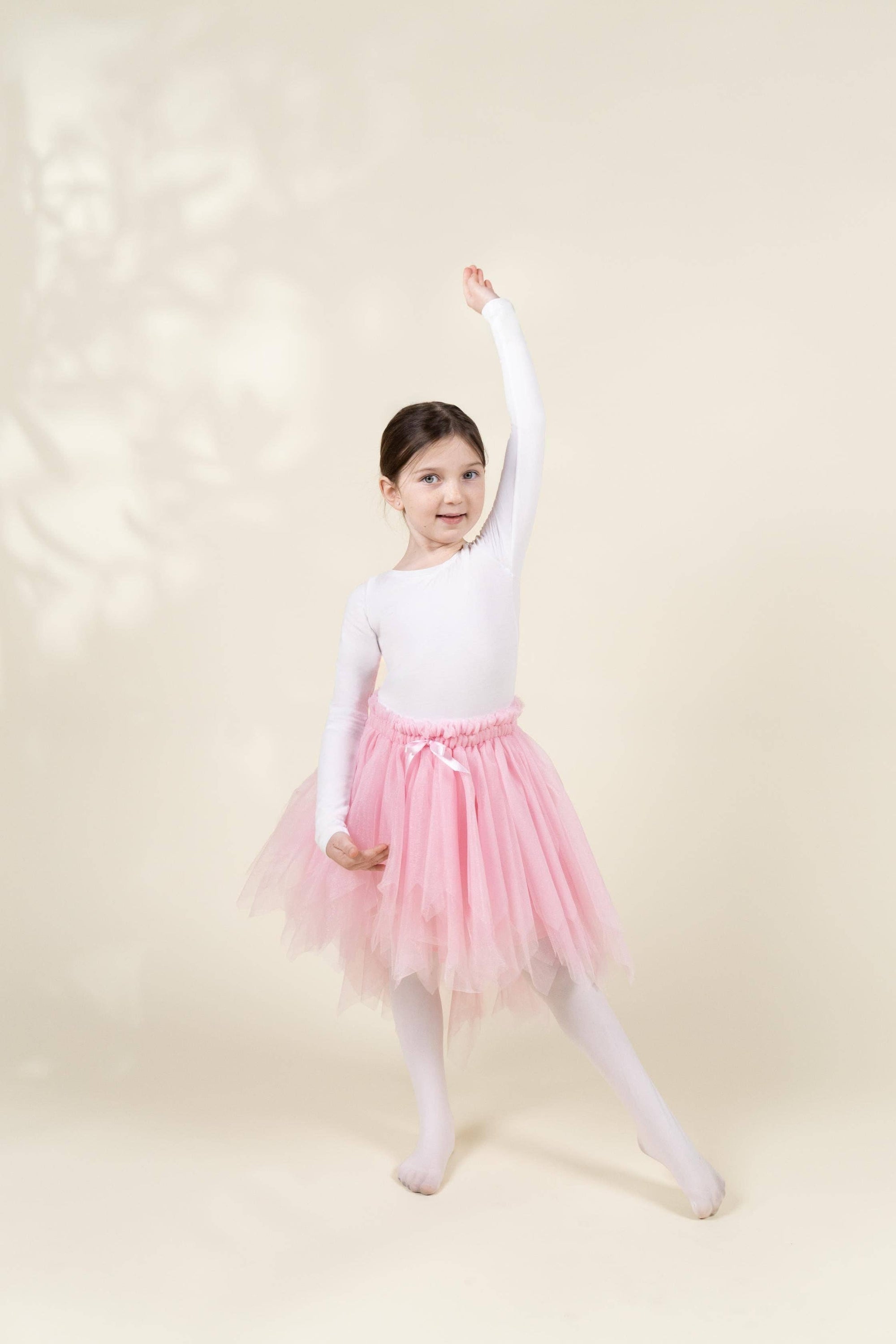 A young girl dancer posing in a white long-sleeve leotard and a pink tulle tutu skirt with white tights, set against a warm beige background with soft leaf shadows.
