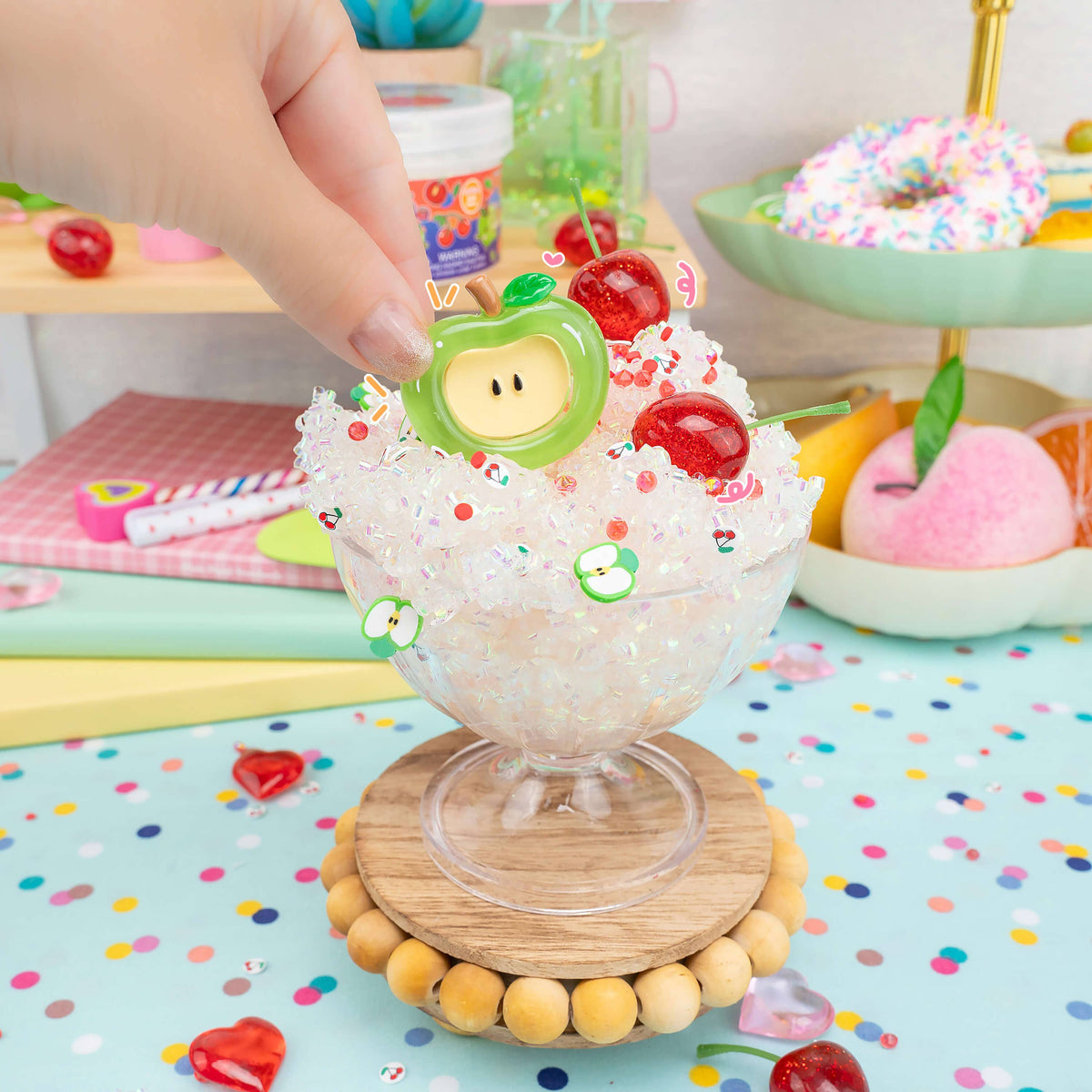 A hand is placing a green apple-shaped charm onto a clear glass dessert cup filled with white glittery material and decorated with red cherries, set on a wooden coaster over a pastel polka-dot surface.