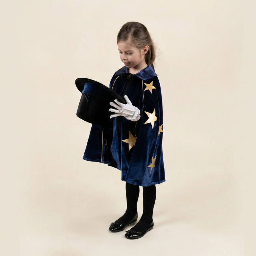 A young girl modeling a dark blue velvet wizard costume cape decorated with gold stars, holding a black magician's hat and wearing white gloves, against a solid beige background.