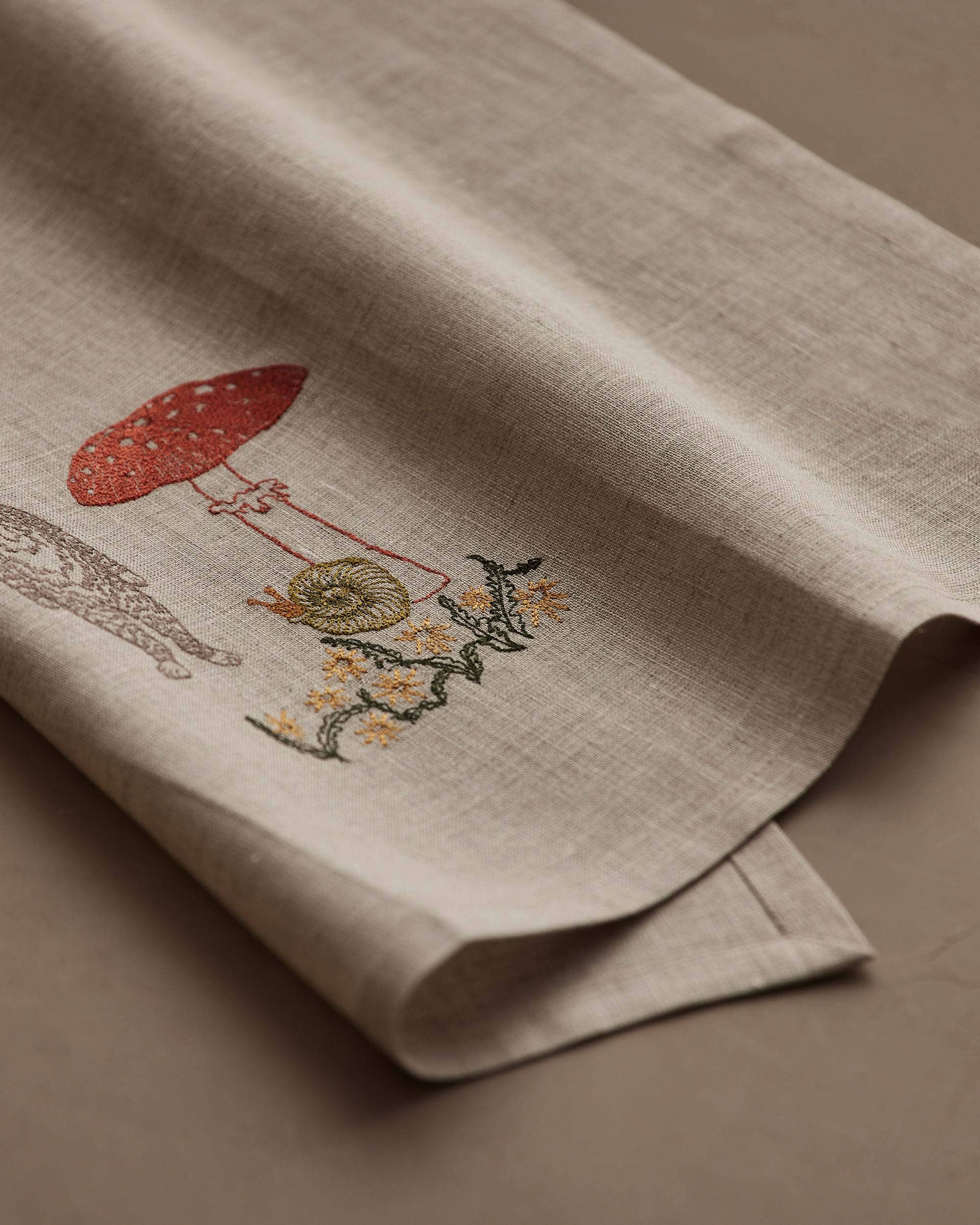 A close-up of a beige linen textile, likely a napkin or table linen, featuring delicate embroidered details of a red mushroom, a snail, and small yellow flowers on a neutral background.