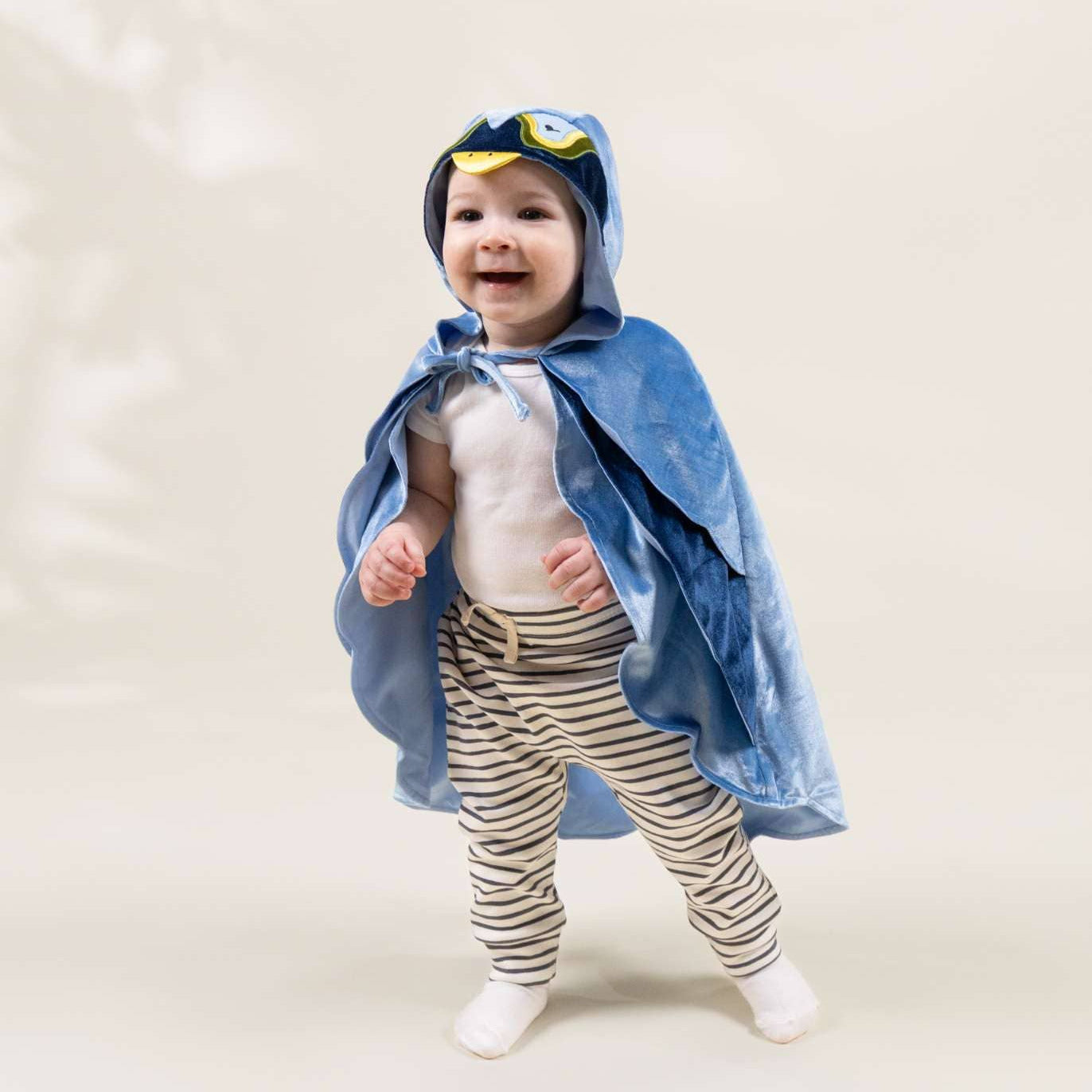 A baby wearing a blue hooded cape with yellow goggles on the hood, a white top, and beige-and-white striped pants, standing against a plain light beige studio background.