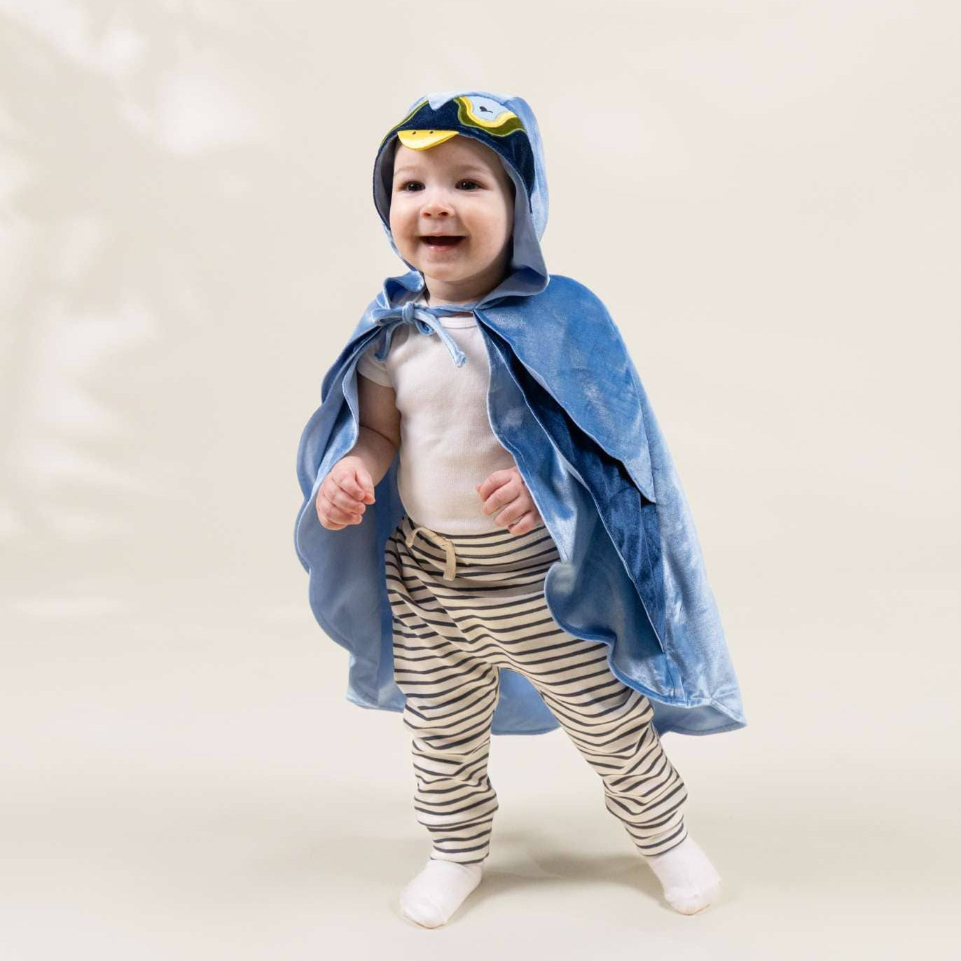 A baby wearing a blue hooded cape with yellow goggles on the hood, a white top, and beige-and-white striped pants, standing against a plain light beige studio background.