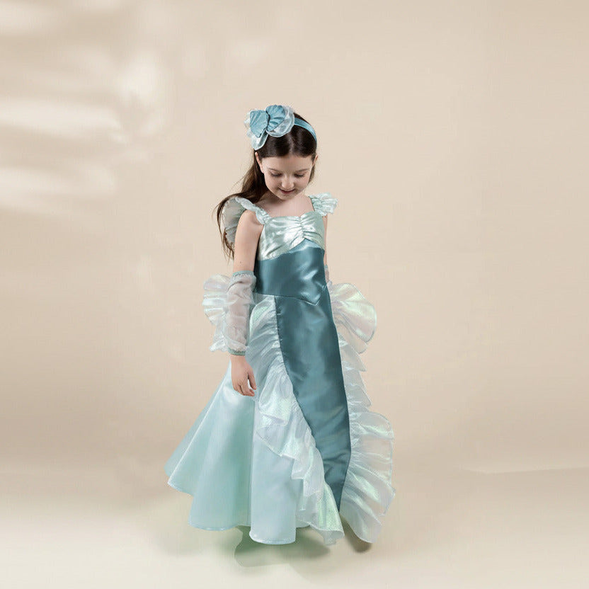 A young girl wearing a light blue and white princess-style dress costume with a matching headband, standing against a plain beige studio background.