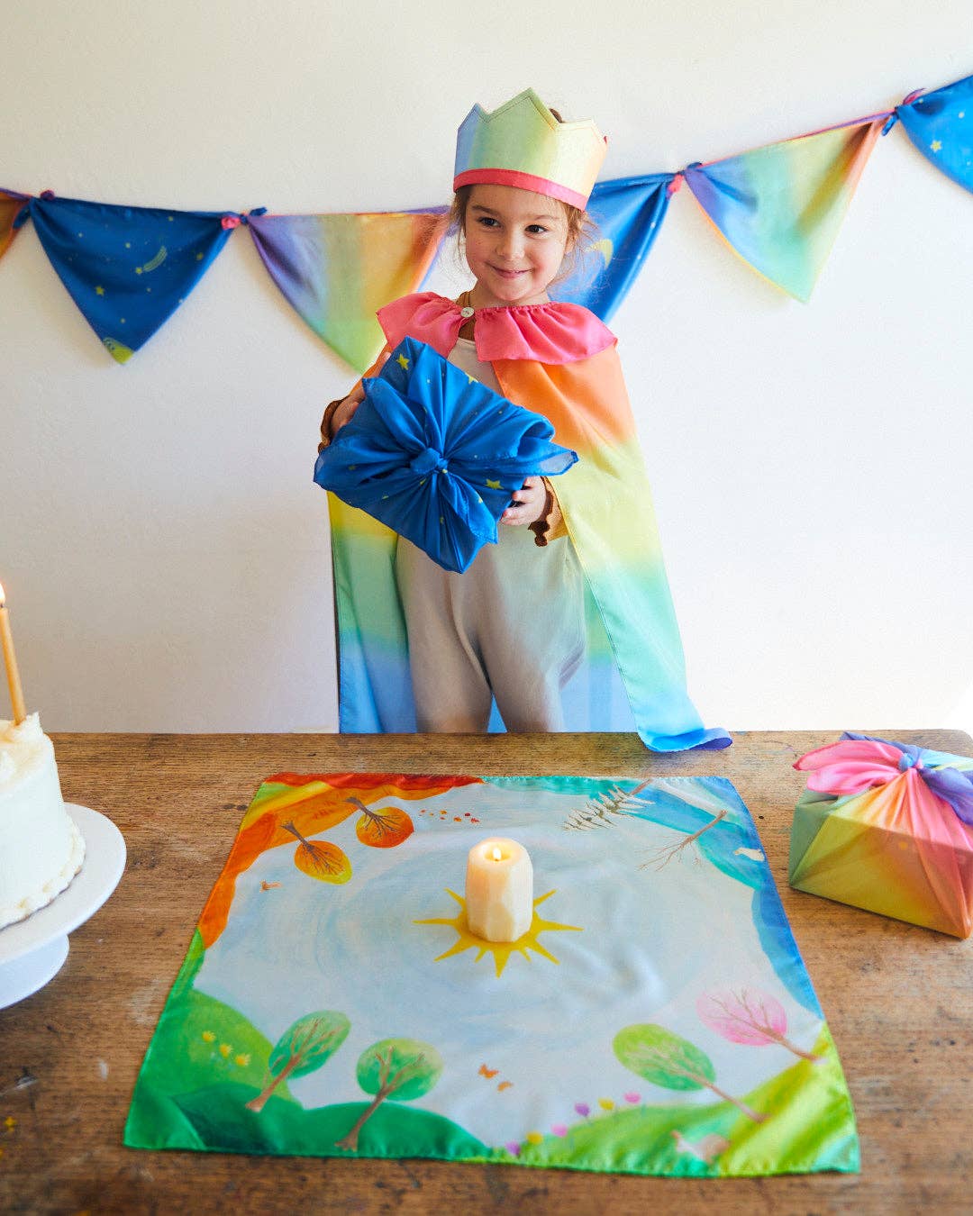 Child in a rainbow-themed costume holding a blue bow, standing behind a table with a colorful placemat and candle.