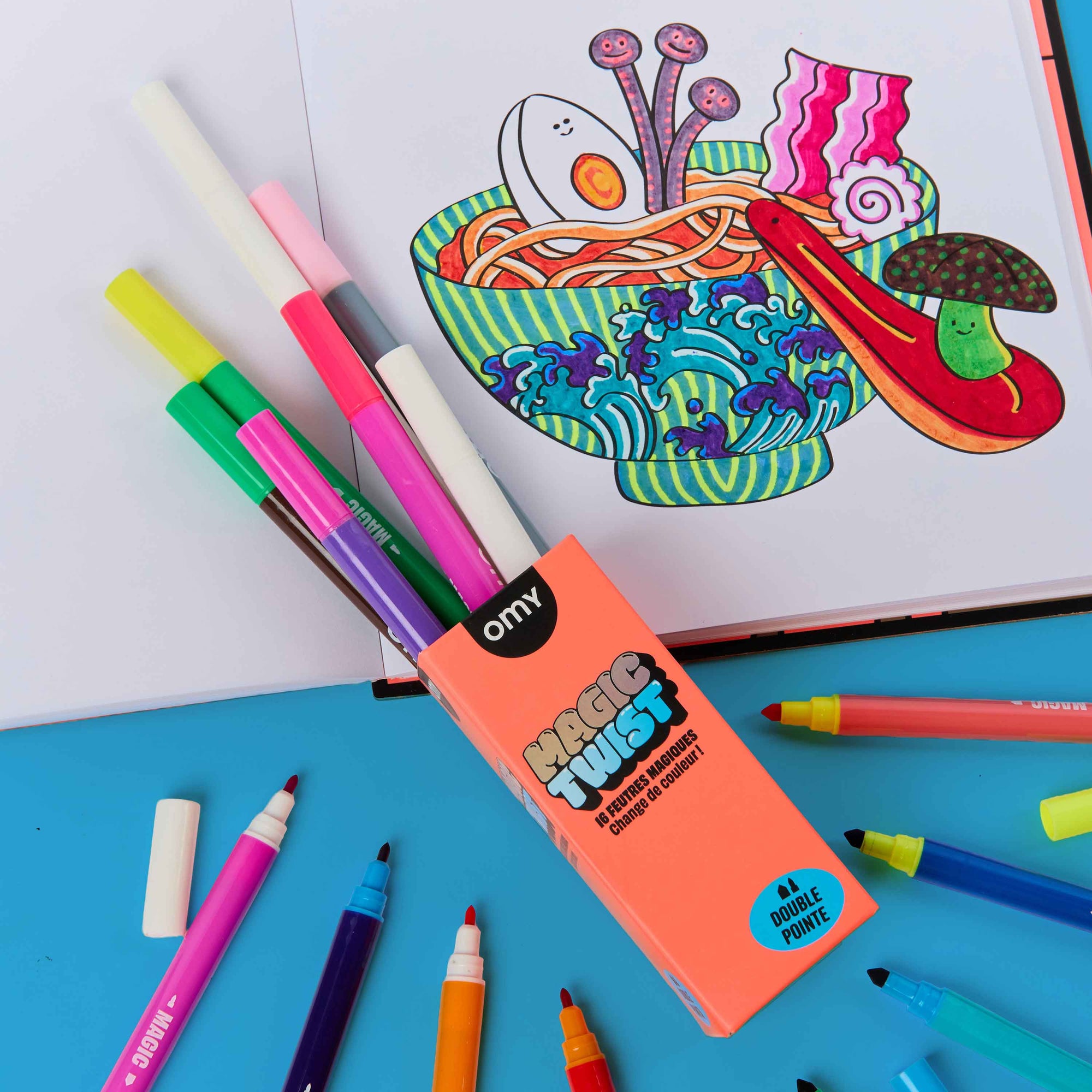 Colorful markers with an open coloring book featuring a bowl of noodles on a blue surface.