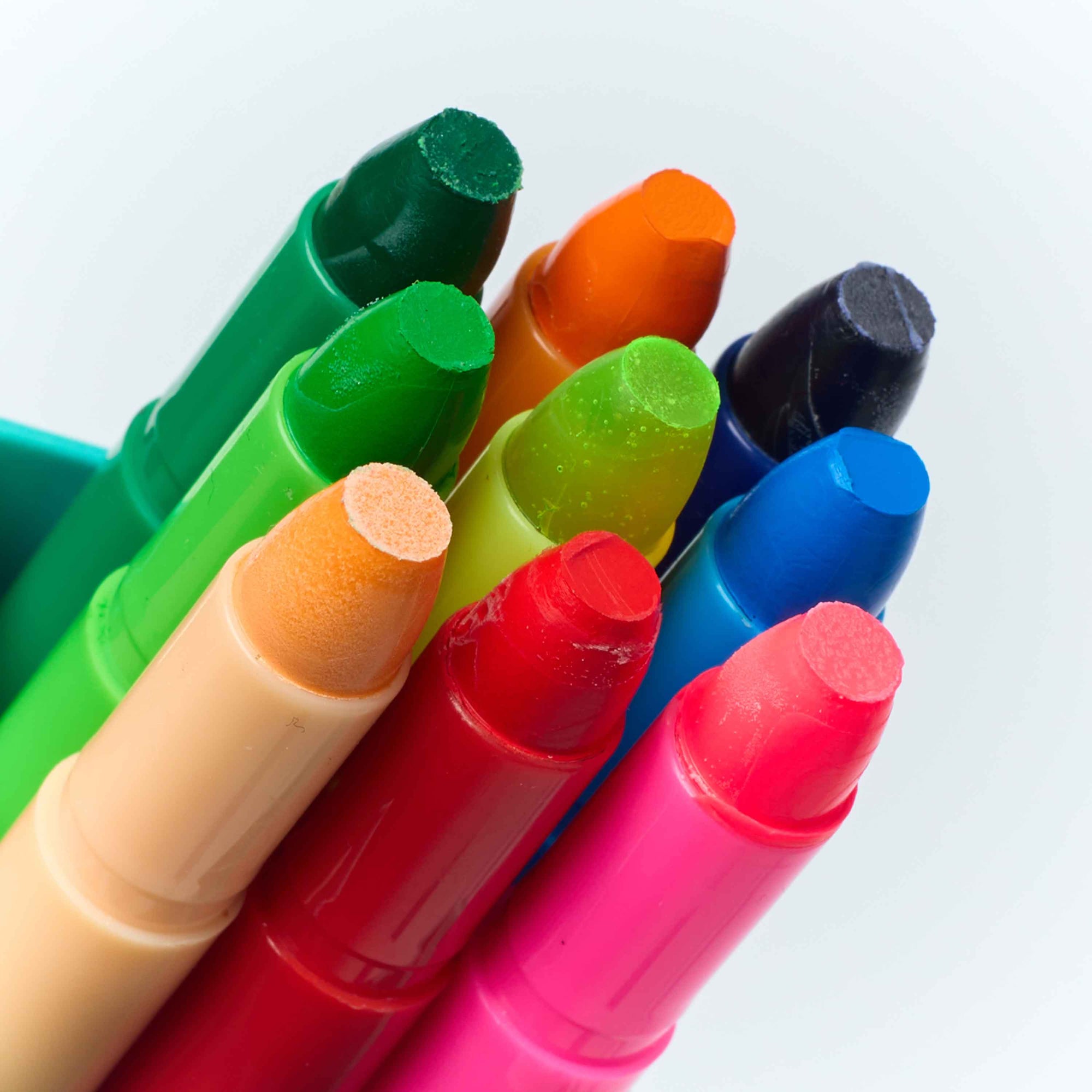 A close-up of multiple crayon-like color sticks in assorted bright colors, including green, orange, blue, pink, red, and beige, arranged together against a light background.