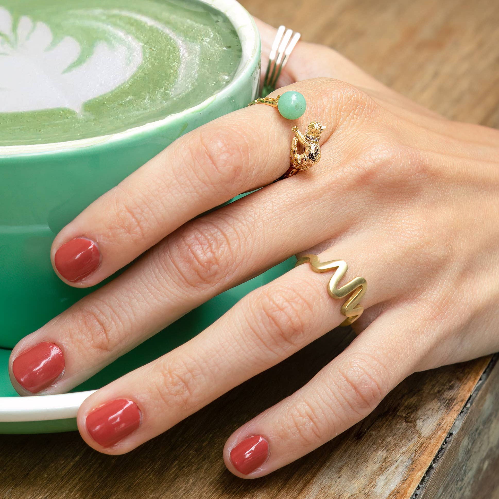 Hand with multiple rings on a wooden surface next to a green cup of coffee.