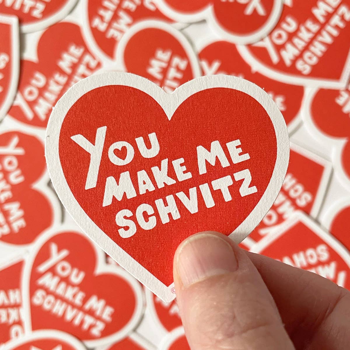 Heart-shaped sticker with 'You Make Me Schvitz' text held by a hand against a background of similar stickers.