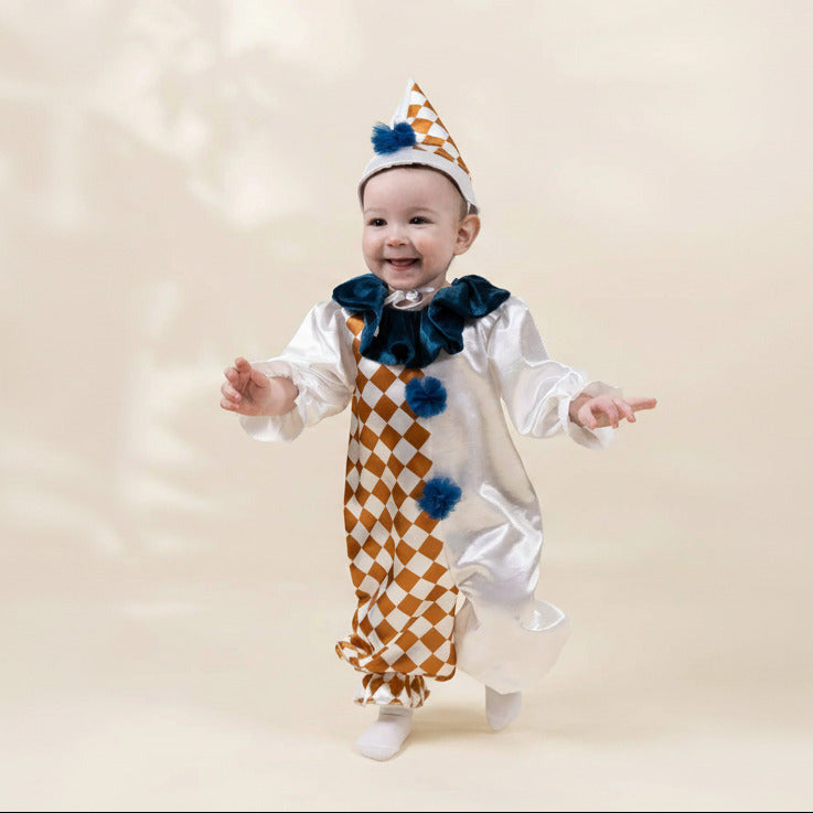 A baby wearing a white and gold checkered clown costume with a dark blue ruffled collar, blue pom-poms, and a matching pointed hat with blue pom-poms, standing against a warm beige background.