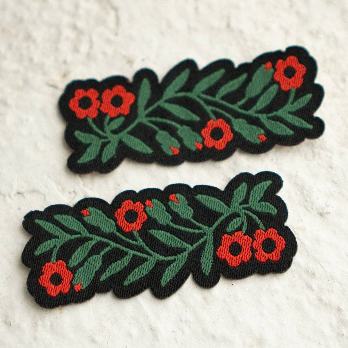 Two embroidered floral hair clips with green leaves and red flowers on a black background, displayed on a textured off-white surface.