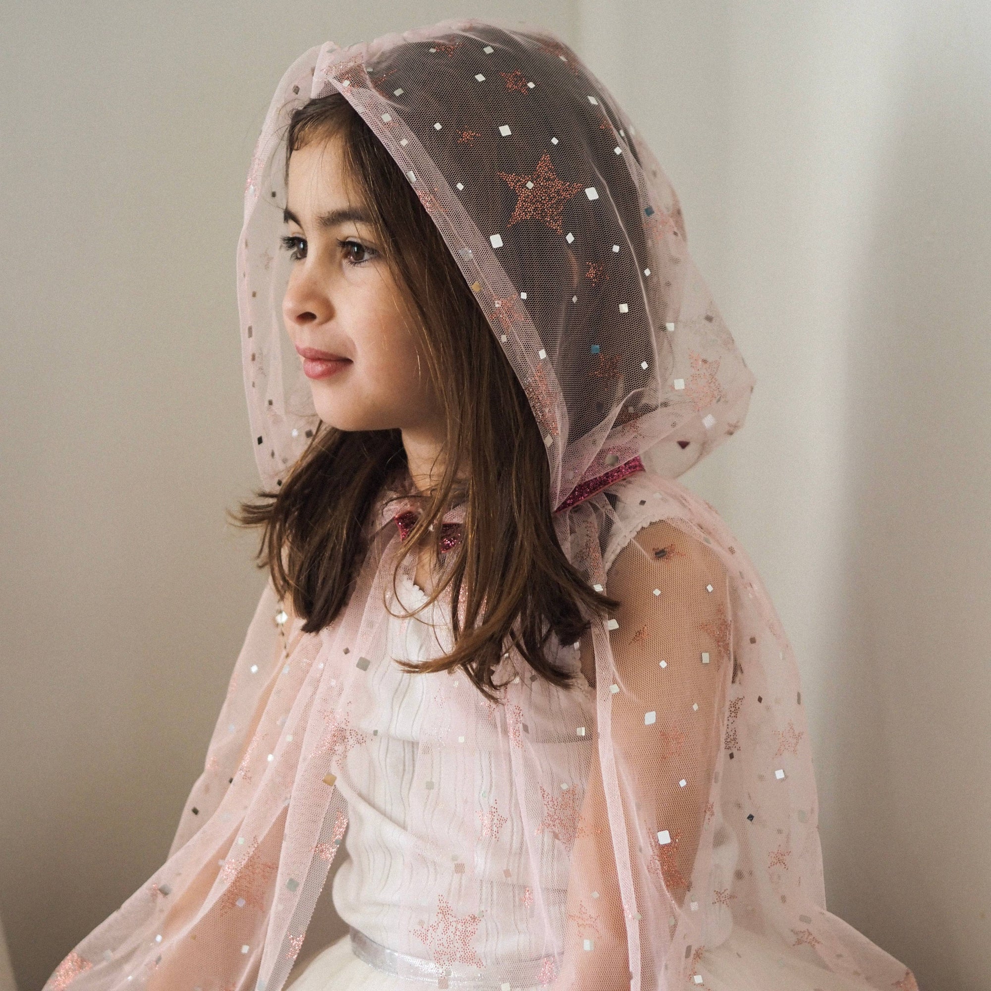 Young girl wearing a white dress with a sheer pink hood against a plain background