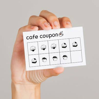 Hand holding a cafe coupon with cupcake illustrations on a gray background