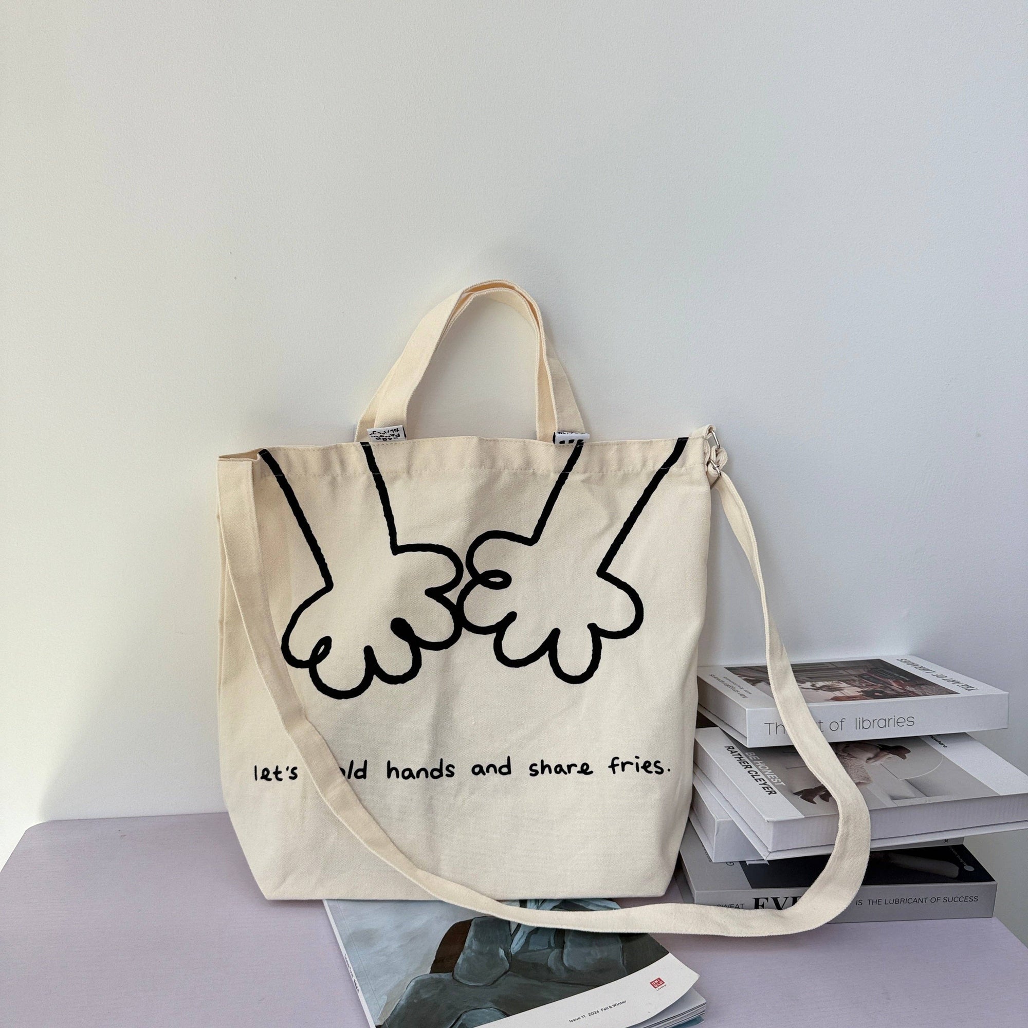 Beige tote bag with black hand design and text on a light gray surface with books.
