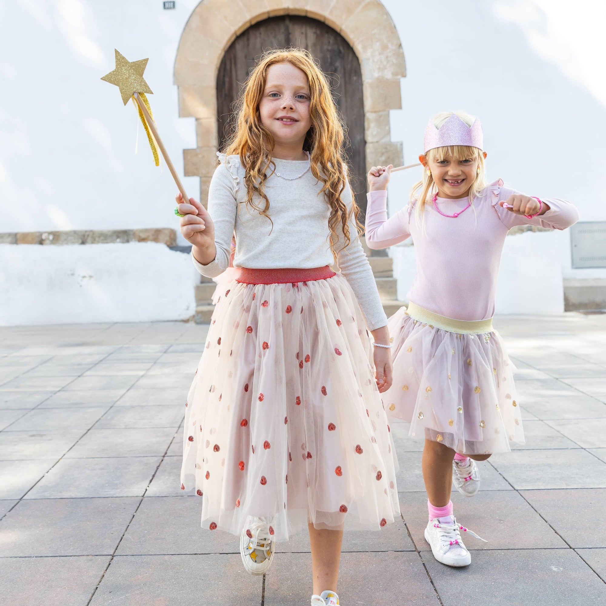 Two children in costumes standing on a tiled pavement with a decorative archway in the background.