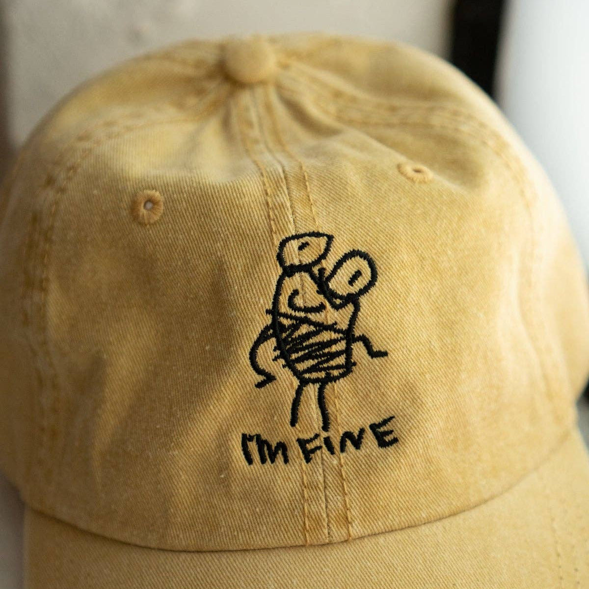 A yellow washed cotton baseball cap featuring a black embroidered stick figure holding a heart and the text 'I'M FINE' underneath, set against a neutral light background.