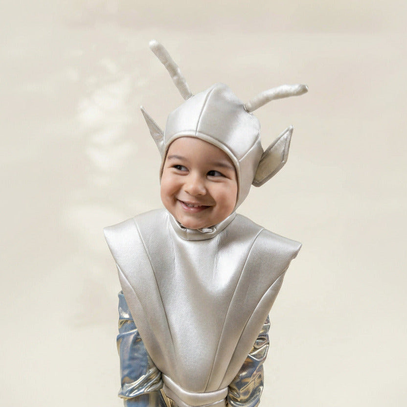 A smiling young child wearing a shiny silver sci-fi alien costume with a helmet featuring two long antennae and pointed ear-like side pieces, against a soft light beige background.