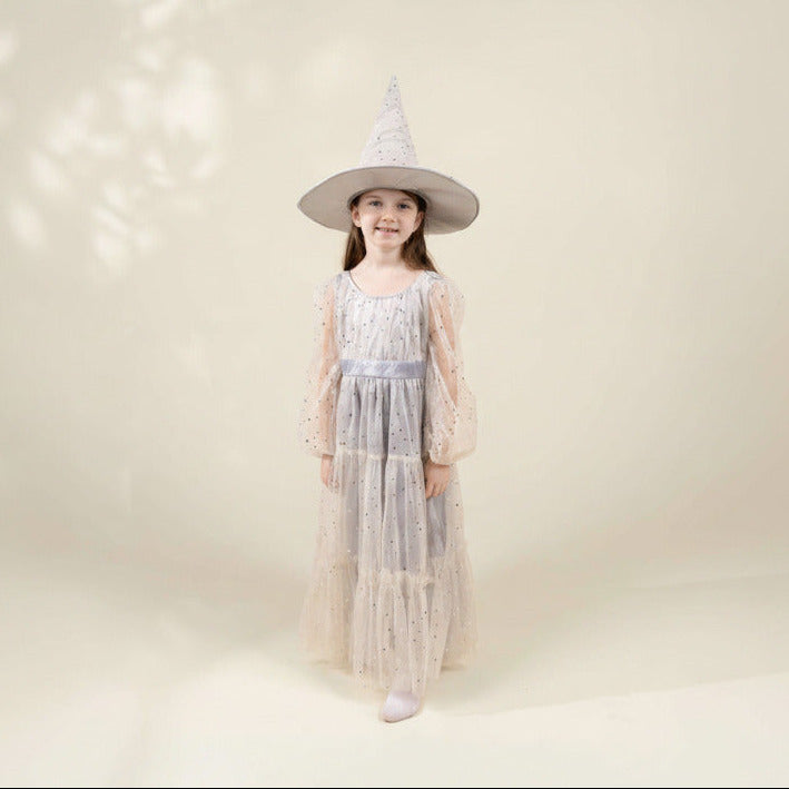 A young girl wearing a wizard or witch costume dress and a tall pointed hat, standing against a warm cream studio backdrop with soft leaf shadows.